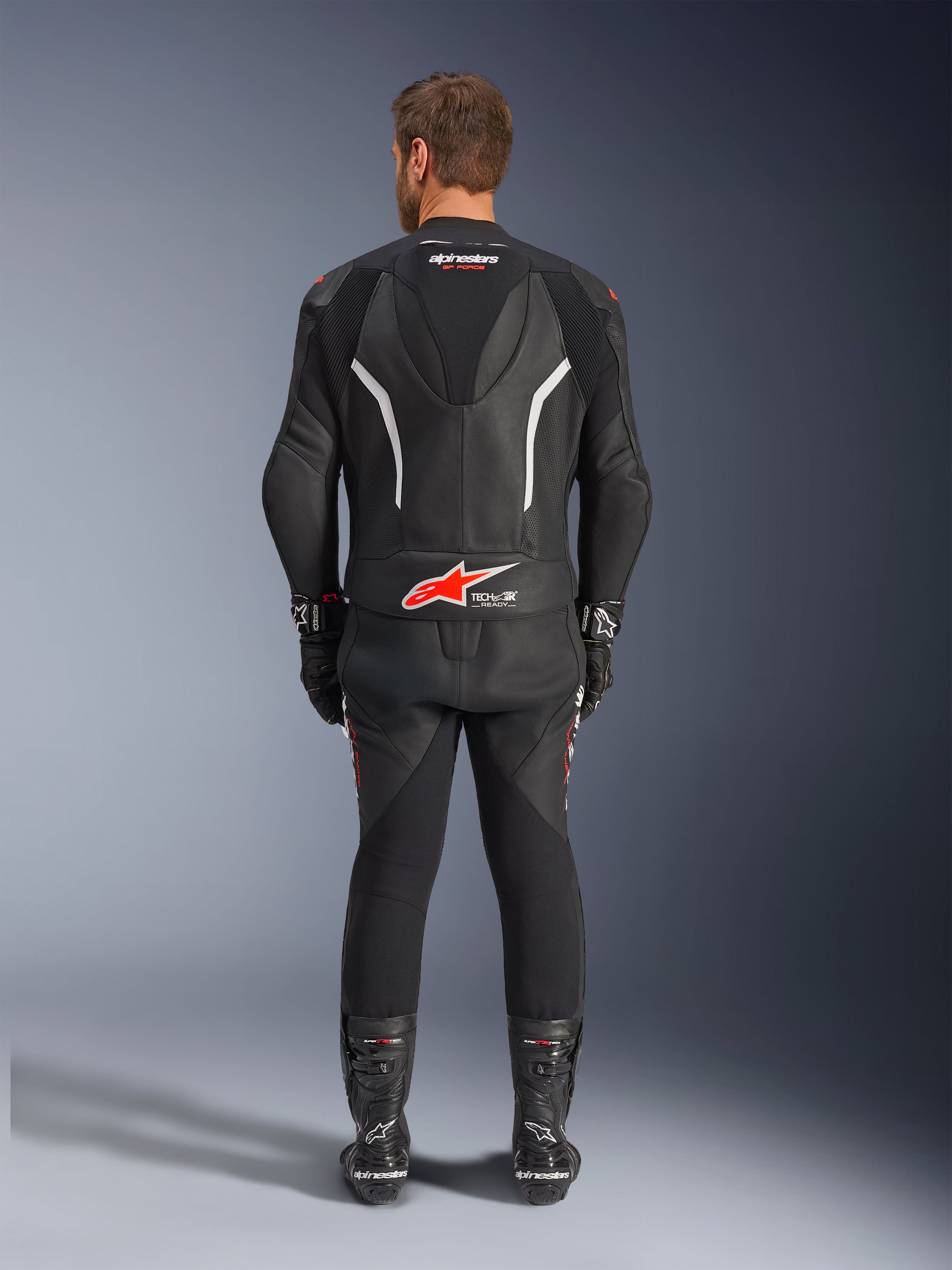 Vista trasera de un piloto con el traje de cuero de 2 piezas Alpinestars GP Force V2, negro con detalles en blanco y rojo, con logotipos prominentes de Alpinestars y Tech-Air Ready en la espalda, combinado con guantes y botas de carreras negros a juego.