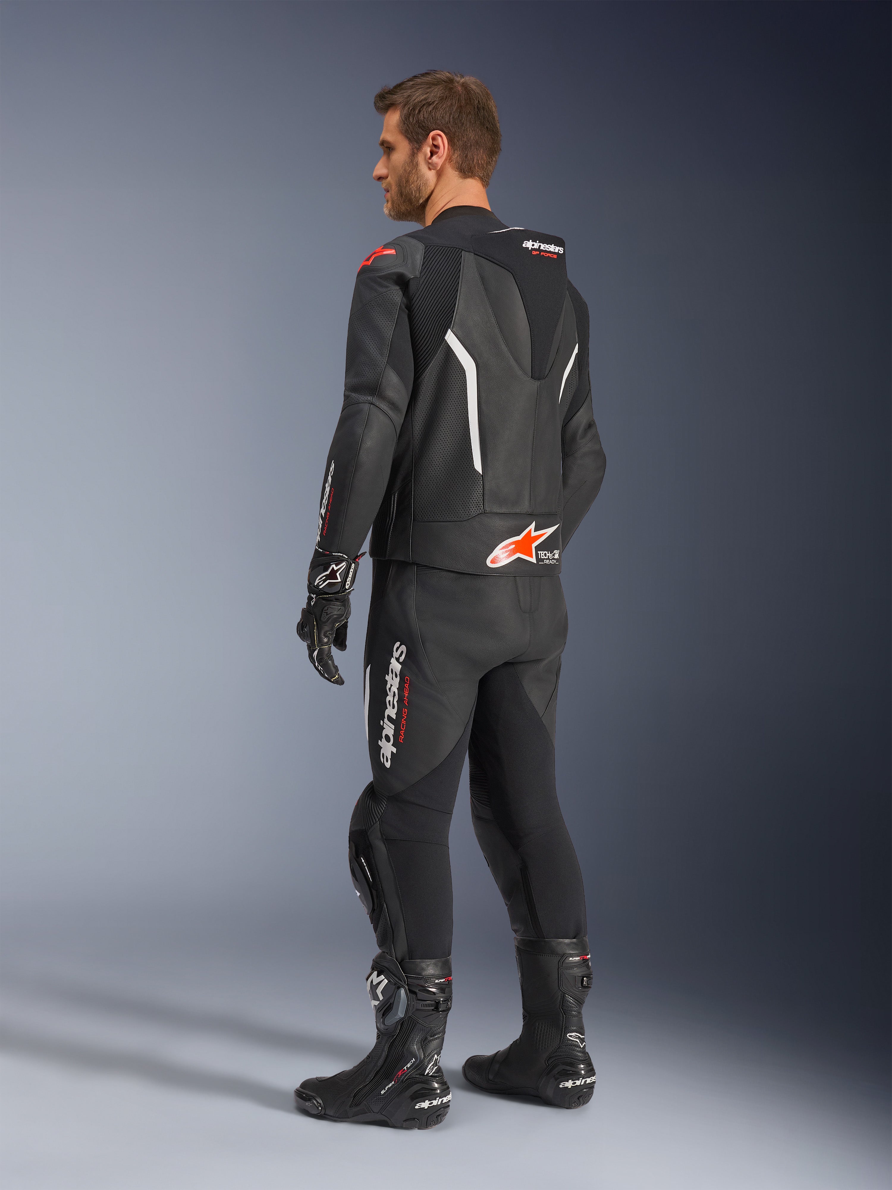 Traje de cuero de 2 piezas Alpinestars GP Force V2, negro con detalles en blanco y rojo flúor, vista trasera de tres cuartos sobre modelo, con joroba aerodinámica, paneles de cuero perforado y logotipos prominentes de Alpinestars, usado con botas y guantes de carreras negros.