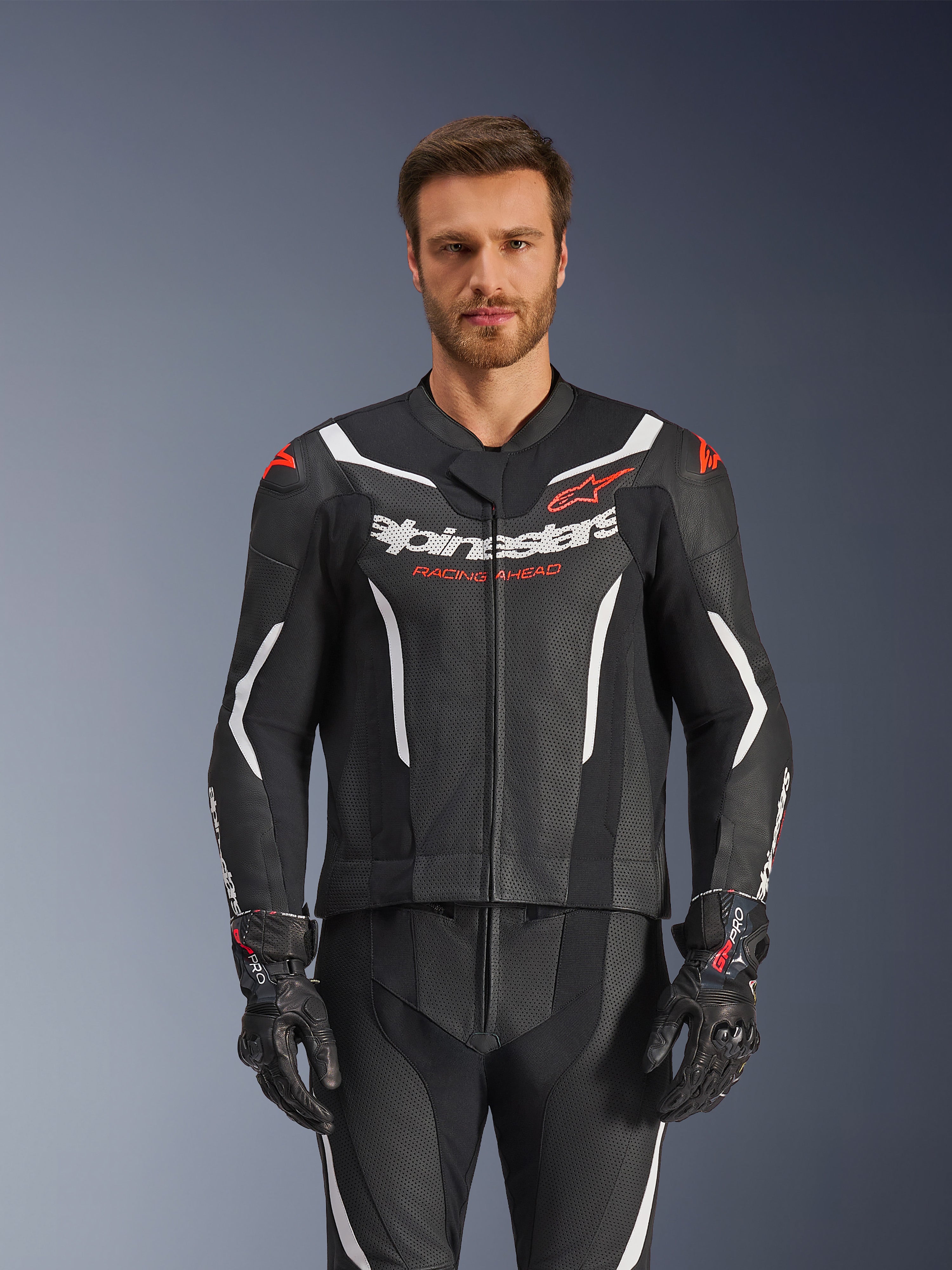 Traje de cuero Alpinestars GP Force V2 de 2 piezas, negro, blanco y rojo fluo, vista frontal de modelo masculino usando el traje de cuero perforado con guantes negros a juego, con detalles blancos angulares y marca Alpinestars en el pecho