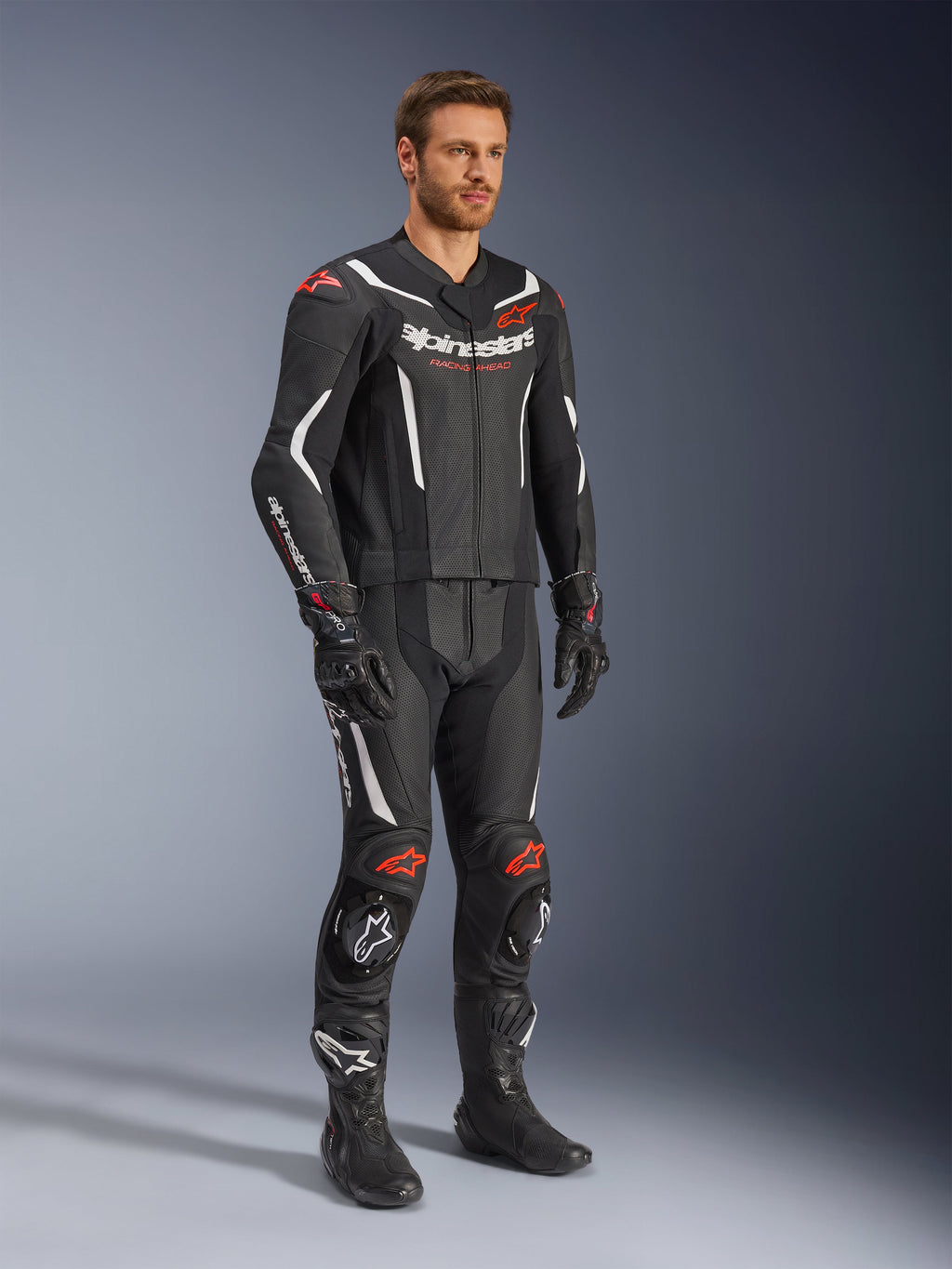 Alpinestars GP Force V2 2-Piece Leather Suit, negro, blanco y rojo flúor, usado por un modelo con botas y guantes de carreras negros, con paneles de cuero perforado, acentos aerodinámicos blancos y logotipos en rojo flúor en hombros y rodillas, equipo profesional de carreras de motociclismo