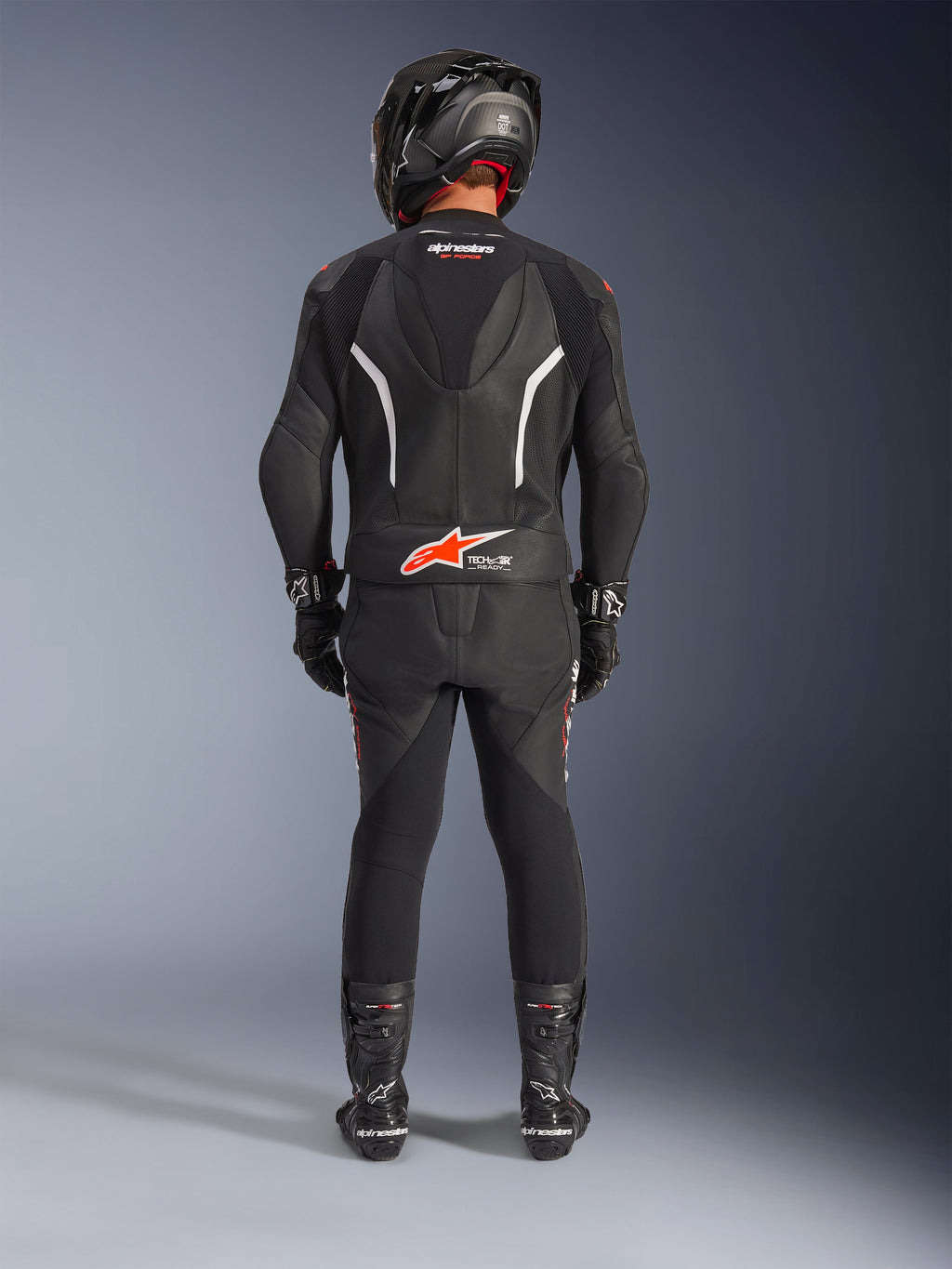 Motociclista visto desde atrás con el traje de cuero de 2 piezas Alpinestars GP Force V2, negro con acentos en blanco y rojo flúor, que incluye joroba aerodinámica y logo Tech-Air Ready, completado con casco de carreras negro, guantes y botas.