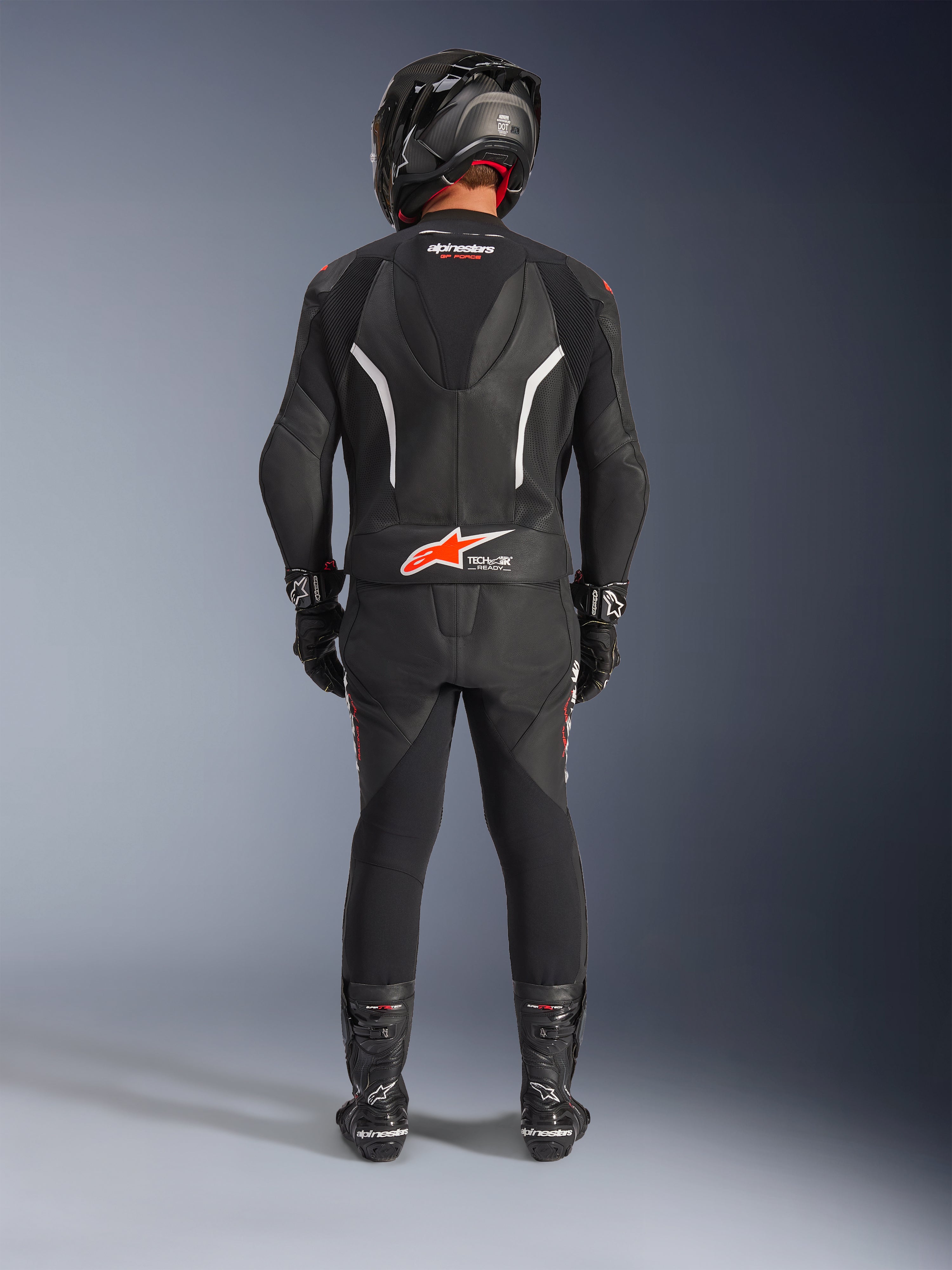 Motociclista visto desde atrás con el traje de cuero de 2 piezas Alpinestars GP Force V2, negro con acentos en blanco y rojo flúor, que incluye joroba aerodinámica y logo Tech-Air Ready, completado con casco de carreras negro, guantes y botas.