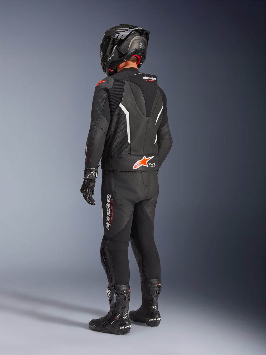 Traje de cuero Alpinestars GP Force V2 de 2 piezas, traje de cuero negro para moto visto desde atrás, usado con casco, guantes y botas negros a juego, con detalles en blanco y rojo, joroba aerodinámica con marca Alpinestars, equipamiento profesional para carreras y conducción deportiva.