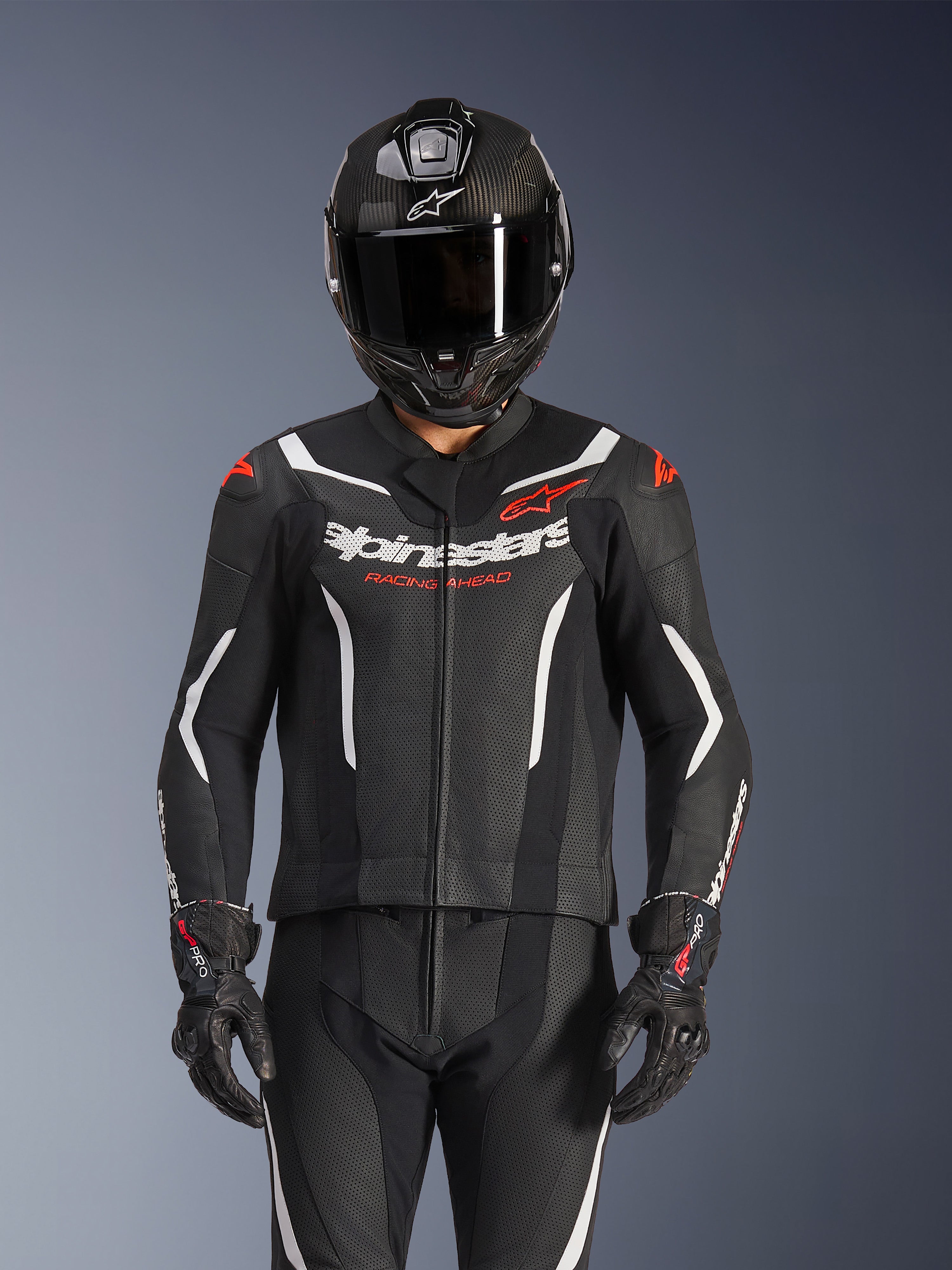 Traje de cuero de 2 piezas Alpinestars GP Force V2, en negro, blanco y rojo flúor, usado por un piloto con casco integral de fibra de carbono y guantes de carreras negros, con paneles de cuero perforado y el logotipo de Alpinestars en el pecho.