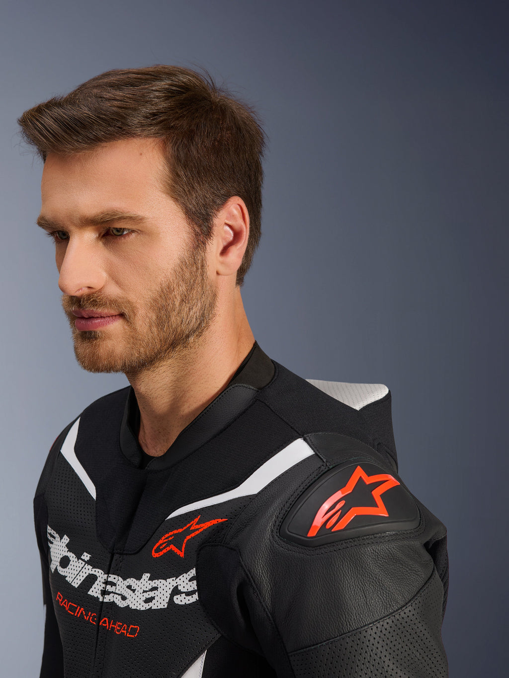 Alpinestars GP Force V2 Leather Suit 2-Piece, primer plano de la chaqueta usada por un modelo masculino, negro, blanco y rojo flúor, con cuero perforado, logotipo de estrella roja en el hombro, joroba aerodinámica y marca Alpinestars en el pecho, equipamiento profesional de carreras de motos