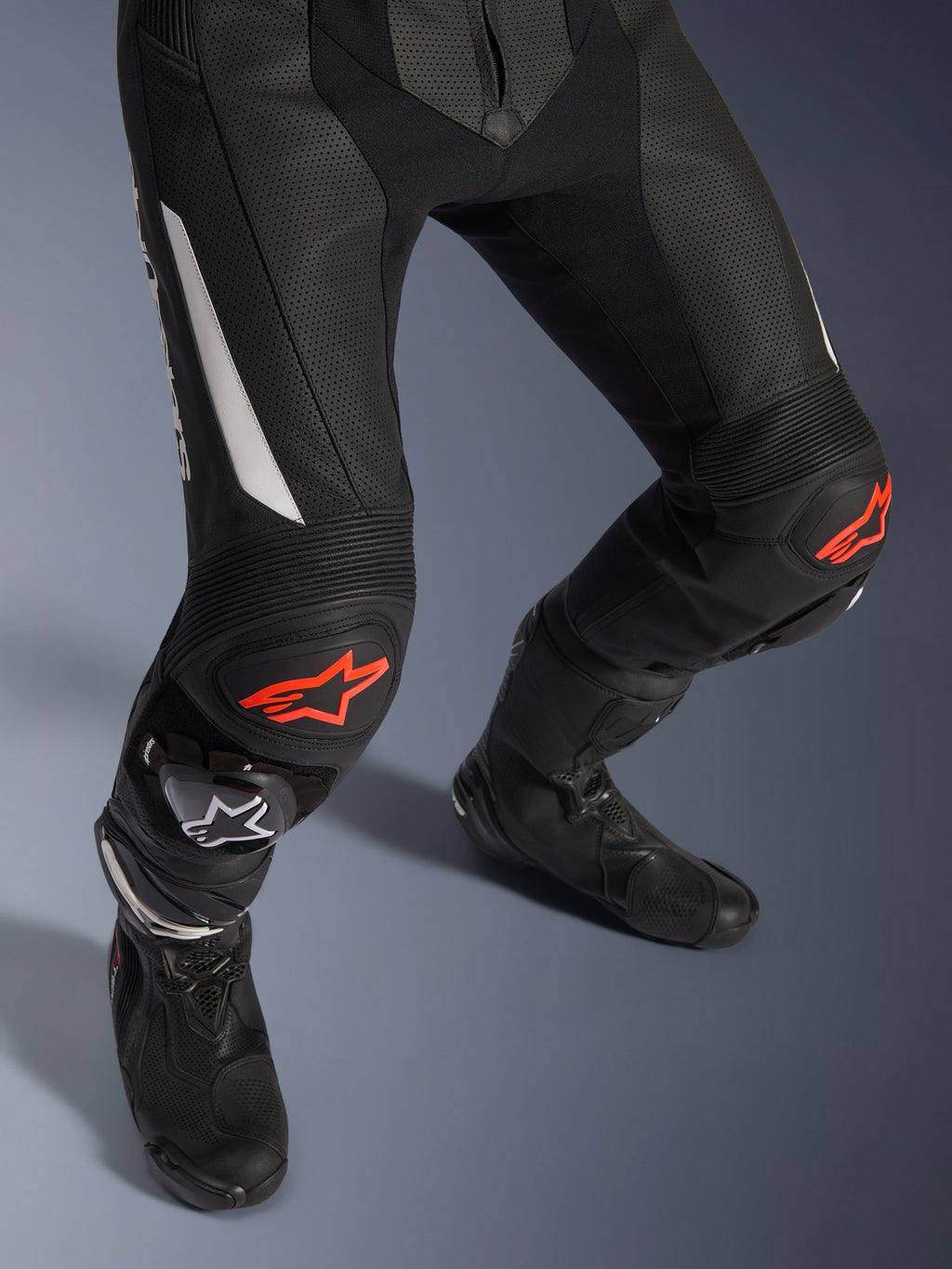 Alpinestars GP Force V2 2-Piece Leather Suit, pantalones de carreras de moto en negro con detalles rojos y blancos, con cuero perforado, paneles elásticos de acordeón sobre las rodillas con logos de estrella roja, usados con botas de moto negras, equipo de carreras de alto rendimiento