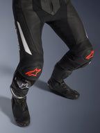 Alpinestars GP Force V2 2-Piece Leather Suit, pantalones de carreras de moto en negro con detalles rojos y blancos, con cuero perforado, paneles elásticos de acordeón sobre las rodillas con logos de estrella roja, usados con botas de moto negras, equipo de carreras de alto rendimiento