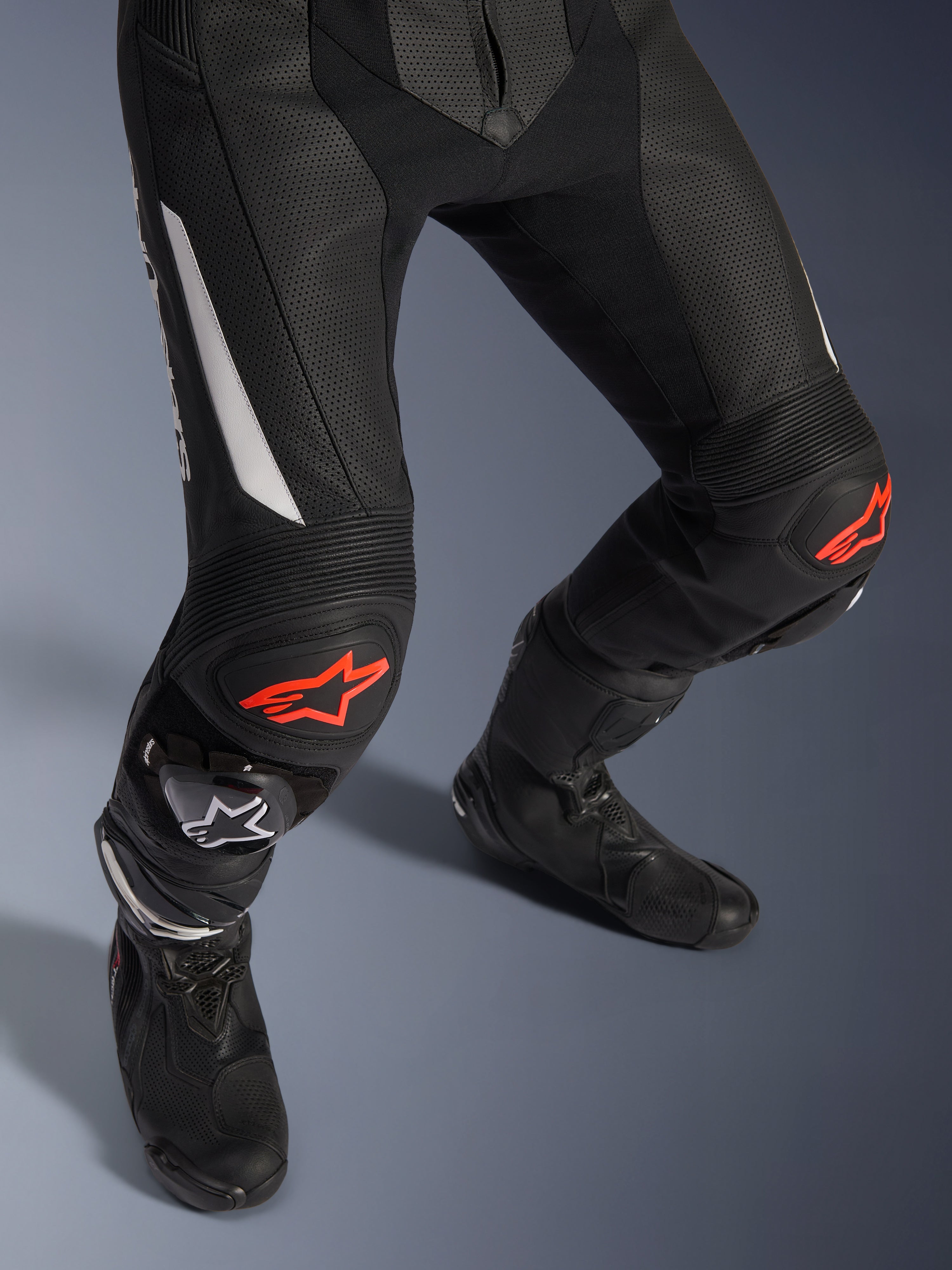 Alpinestars GP Force V2 2-Piece Leather Suit, pantalones de carreras de moto en negro con detalles rojos y blancos, con cuero perforado, paneles elásticos de acordeón sobre las rodillas con logos de estrella roja, usados con botas de moto negras, equipo de carreras de alto rendimiento