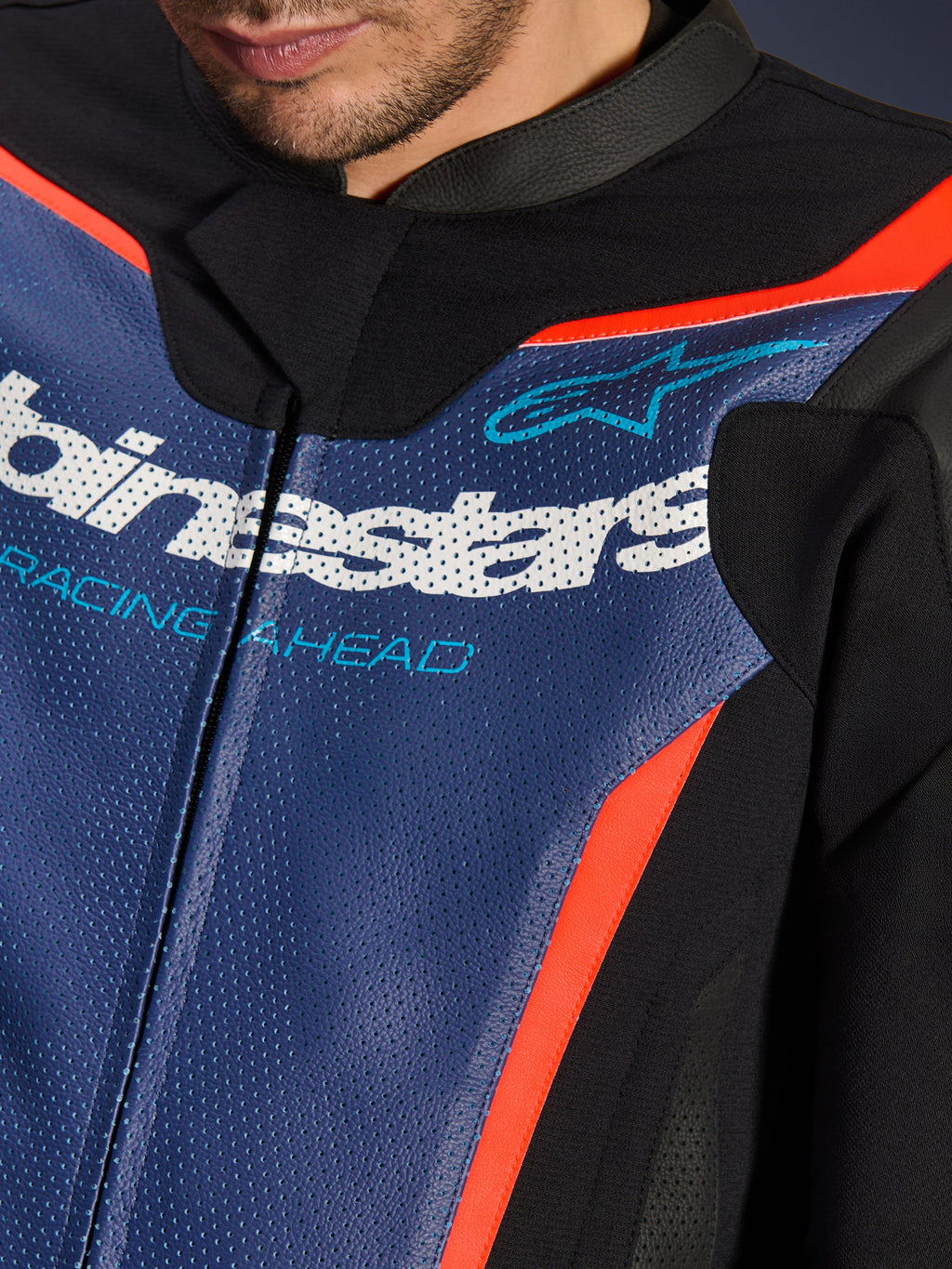 Traje de cuero Alpinestars GP Force V2 de 2 piezas, primer plano de la zona del pecho, cuero azul perforado con logo blanco y texto 'RACING AHEAD', detalles en rojo flúor y paneles negros, equipamiento profesional para conducción deportiva