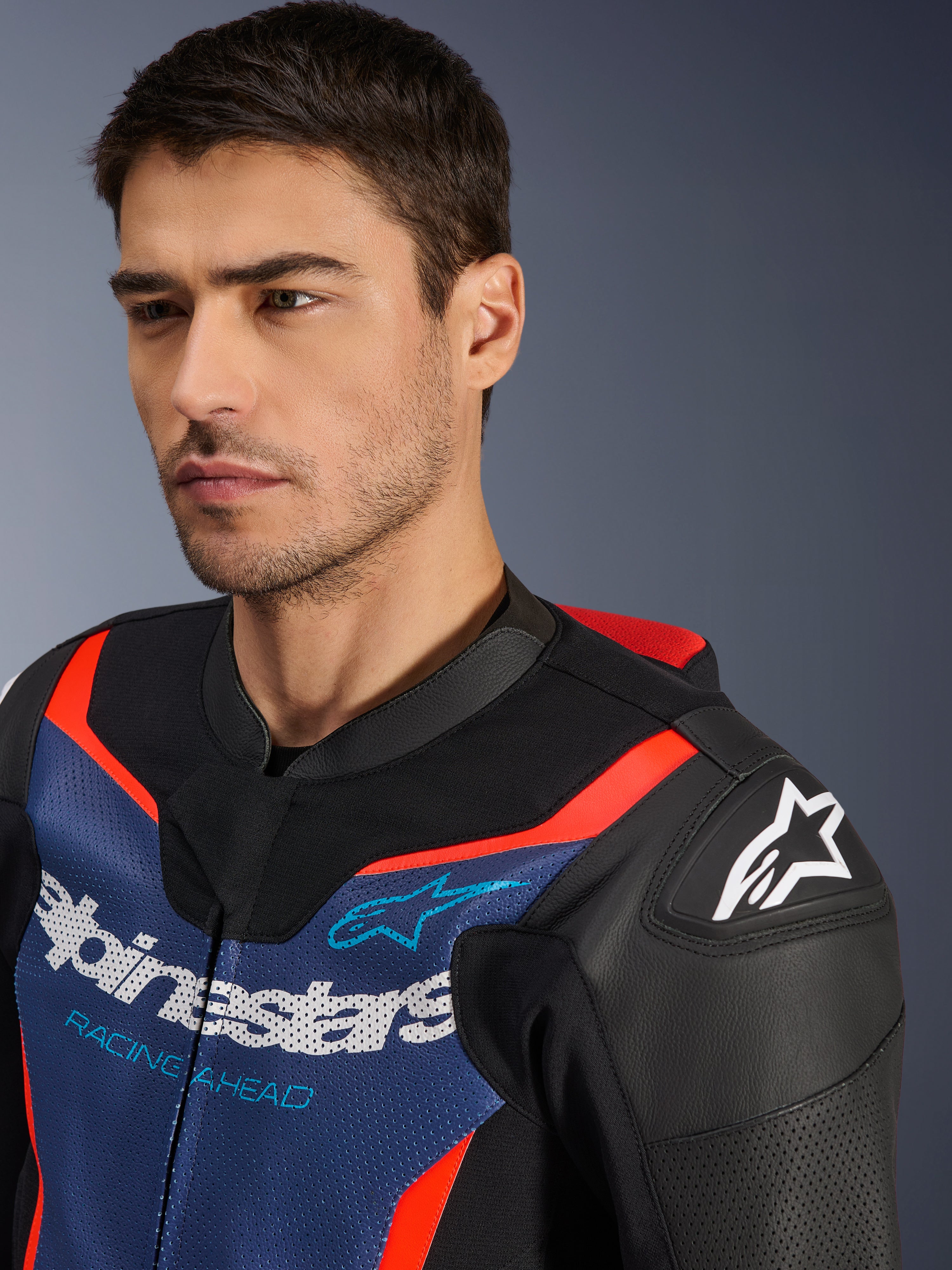 Traje de cuero Alpinestars GP Force V2 de 2 piezas, negro, azul y rojo flúor, primer plano de un modelo con la chaqueta del traje, con paneles de cuero bovino perforado, logotipo de Alpinestars en el hombro y marca 'Racing Ahead' en el pecho, equipo de carreras de nivel profesional