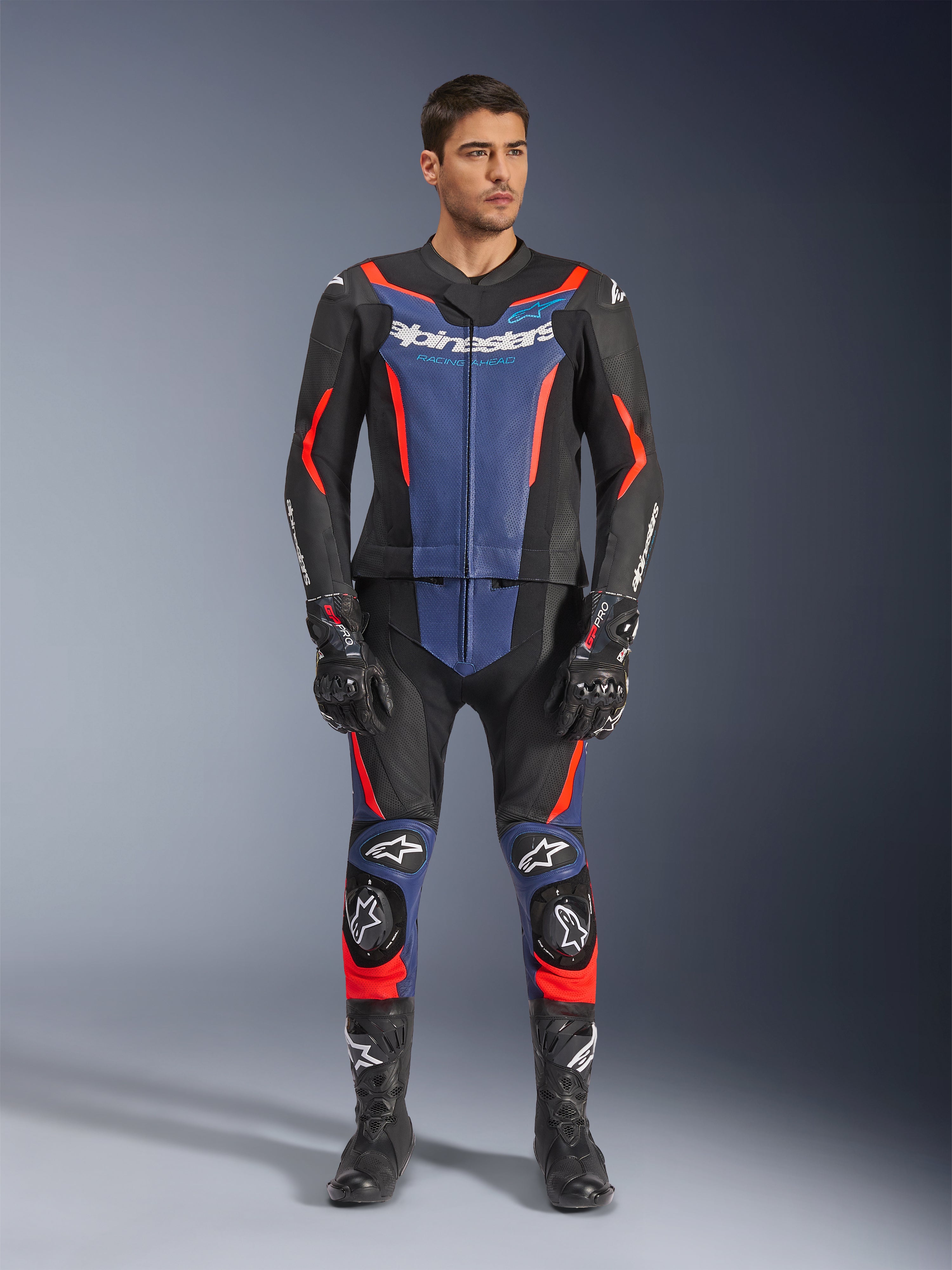 Mono de cuero de 2 piezas Alpinestars GP Force V2 en negro, azul y rojo flúor, con paneles azules en el pecho y la parte inferior de las piernas con vibrantes detalles en rojo fluorescente, vestido por un modelo masculino con botas y guantes de carreras negros, equipamiento de protección profesional para deportes de motor