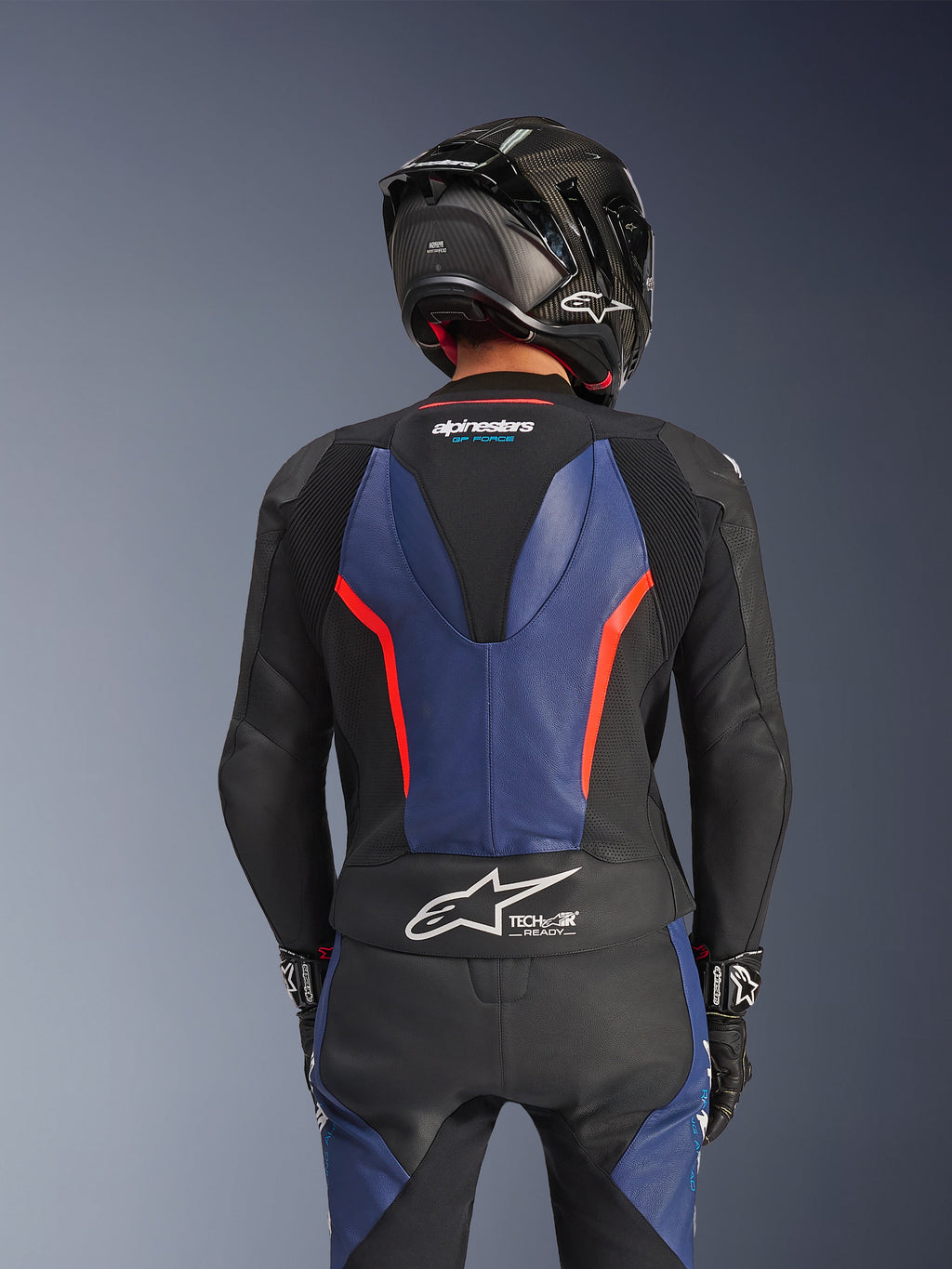 Traje de cuero de dos piezas Alpinestars GP Force V2, color negro, azul y rojo flúor, vista trasera que muestra la joroba aerodinámica y el logotipo Tech-Air Ready, cuero bovino premium con zonas elásticas, usado con un casco de fibra de carbono negro, equipamiento profesional para carreras de motos.
