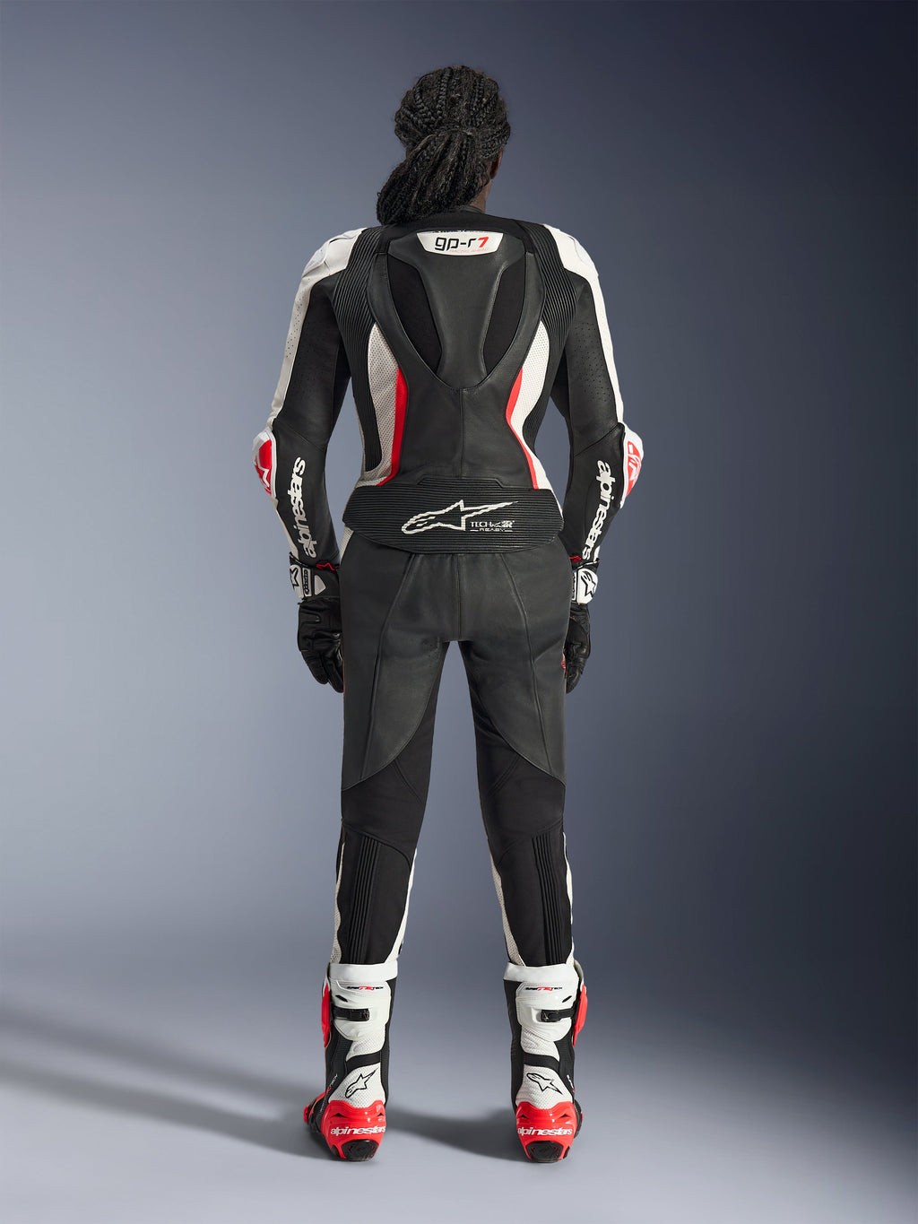 Vista posterior del traje de cuero de 2 piezas Alpinestars Stella GP-R7, negro, blanco y rojo flúor, con una joroba aerodinámica con marca GP-R7 y logotipo Tech-Air Ready, usado con botas y guantes de carreras a juego, equipamiento profesional de motociclismo para mujer