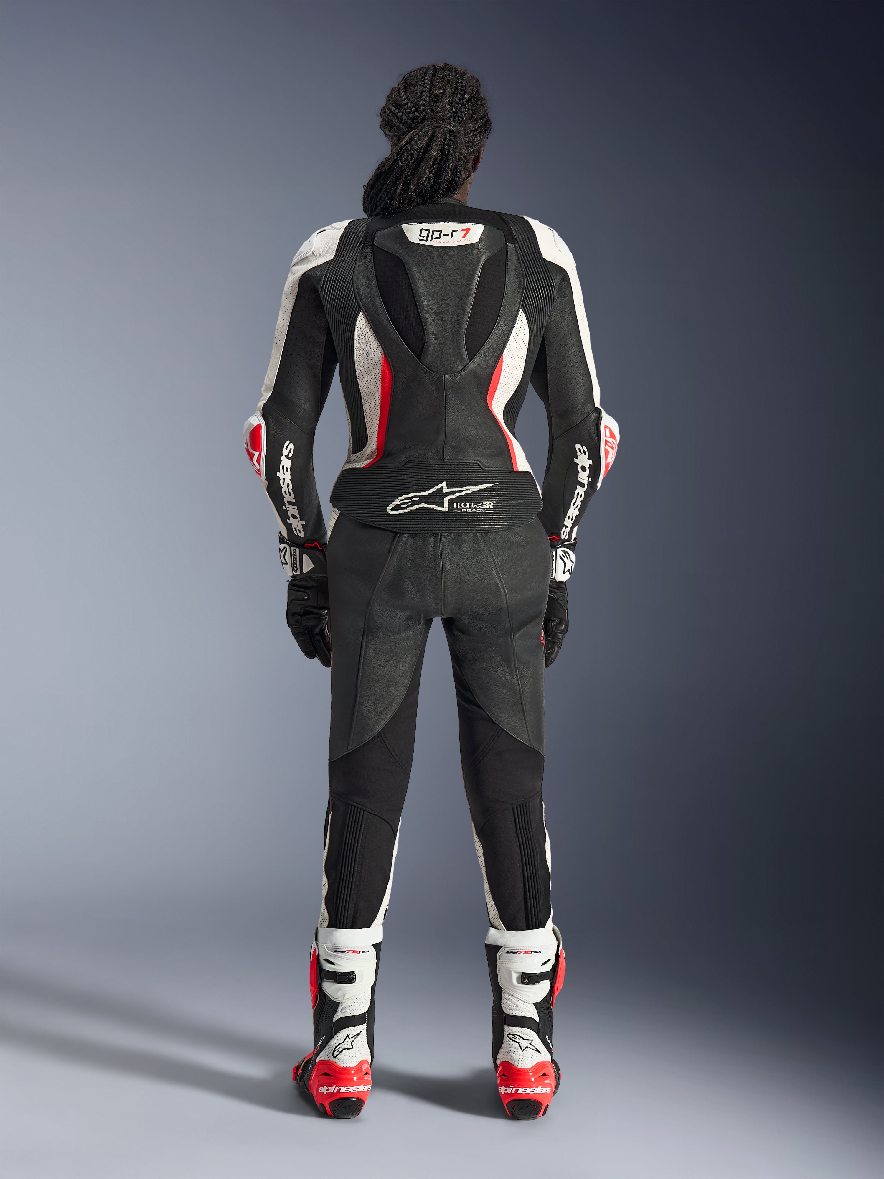 Vista posterior del traje de cuero de 2 piezas Alpinestars Stella GP-R7, negro, blanco y rojo flúor, con una joroba aerodinámica con marca GP-R7 y logotipo Tech-Air Ready, usado con botas y guantes de carreras a juego, equipamiento profesional de motociclismo para mujer