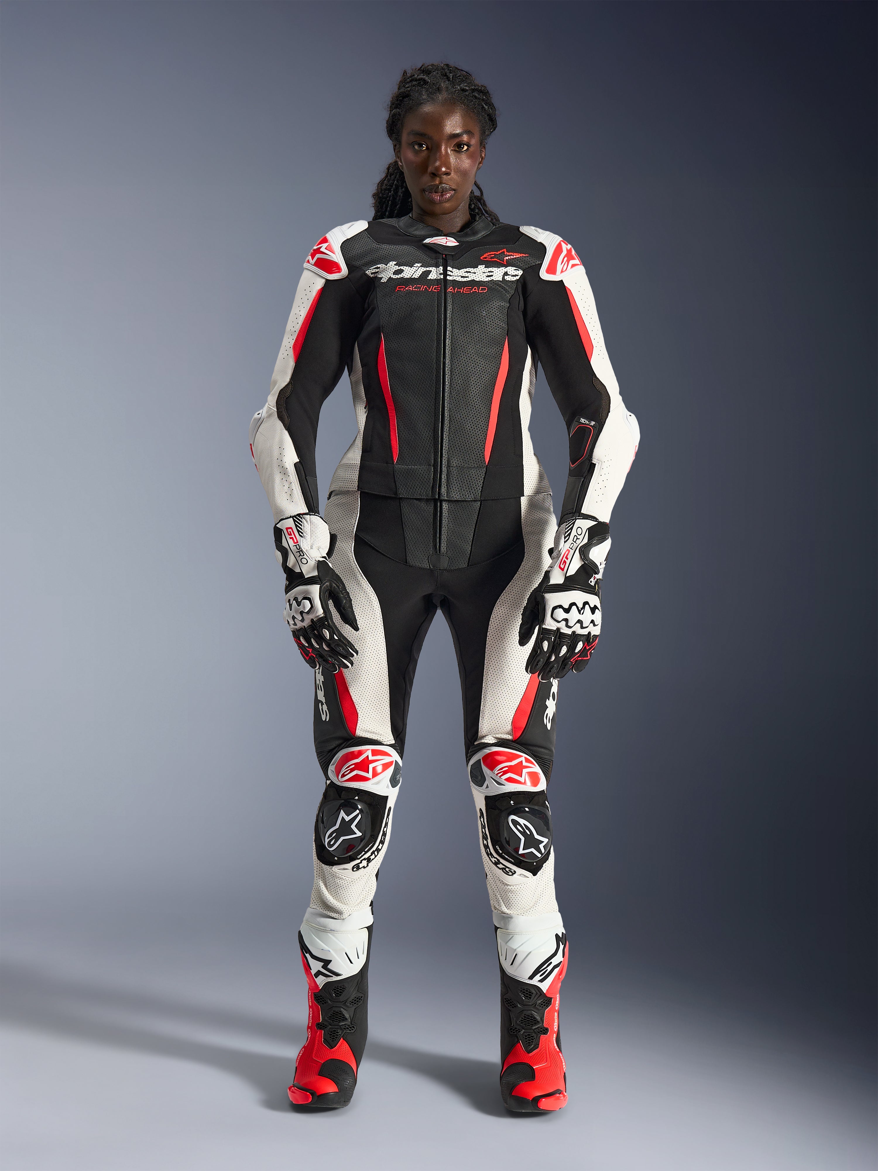 Modelo femenina vistiendo el traje de cuero de 2 piezas Alpinestars Stella GP-R7 en negro, blanco y rojo flúor, completo con guantes y botas de competición a juego, con armadura protectora y paneles de cuero perforados, equipo profesional de carreras de motociclismo