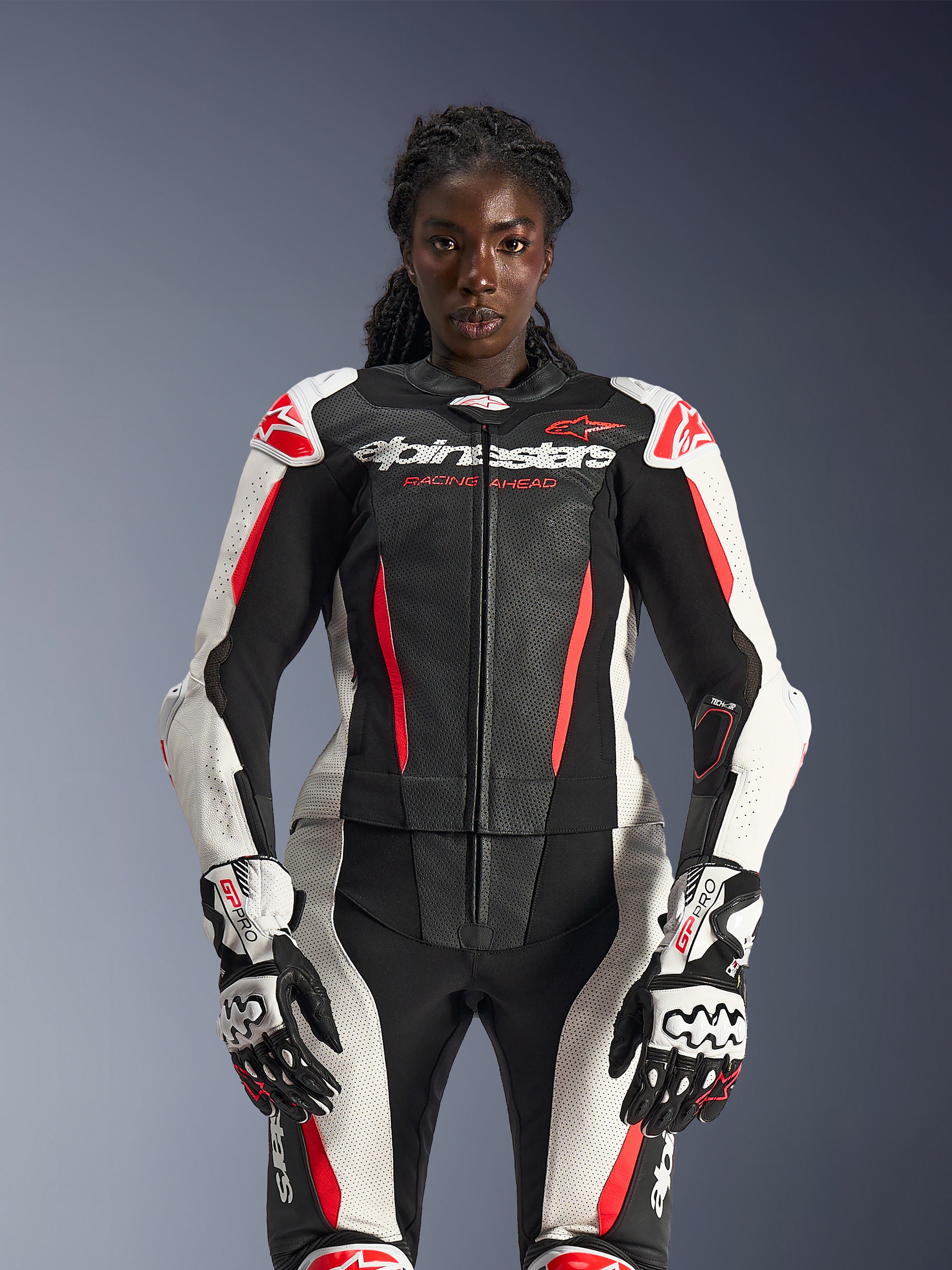 Modelo femenina con el traje de cuero Alpinestars Stella GP-R7 de 2 piezas en negro, blanco y rojo flúor, con paneles de cuero perforados y guantes GP Pro a juego, optimizado anatómicamente para mujeres.