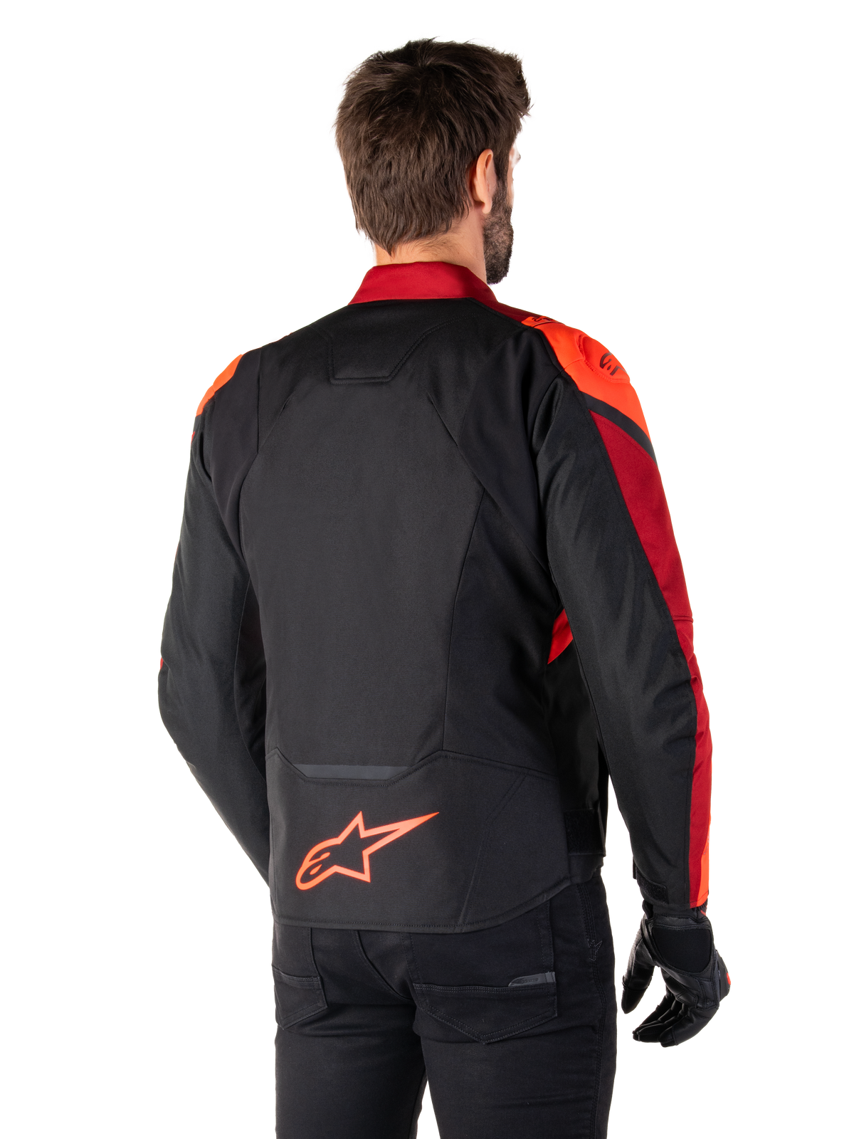 Vista trasera de un hombre con la chaqueta Alpinestars T-Jaws V4 Waterproof en Negro Rojo Fluo Rojo Oscuro, con detalles en rojo vibrante en hombros y mangas, logotipo de Alpinestars en rojo flúor en la parte inferior de la espalda, combinada con pantalones y guantes negros.