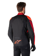 Vista trasera de un hombre con la chaqueta Alpinestars T-Jaws V4 Waterproof en Negro Rojo Fluo Rojo Oscuro, con detalles en rojo vibrante en hombros y mangas, logotipo de Alpinestars en rojo flúor en la parte inferior de la espalda, combinada con pantalones y guantes negros.