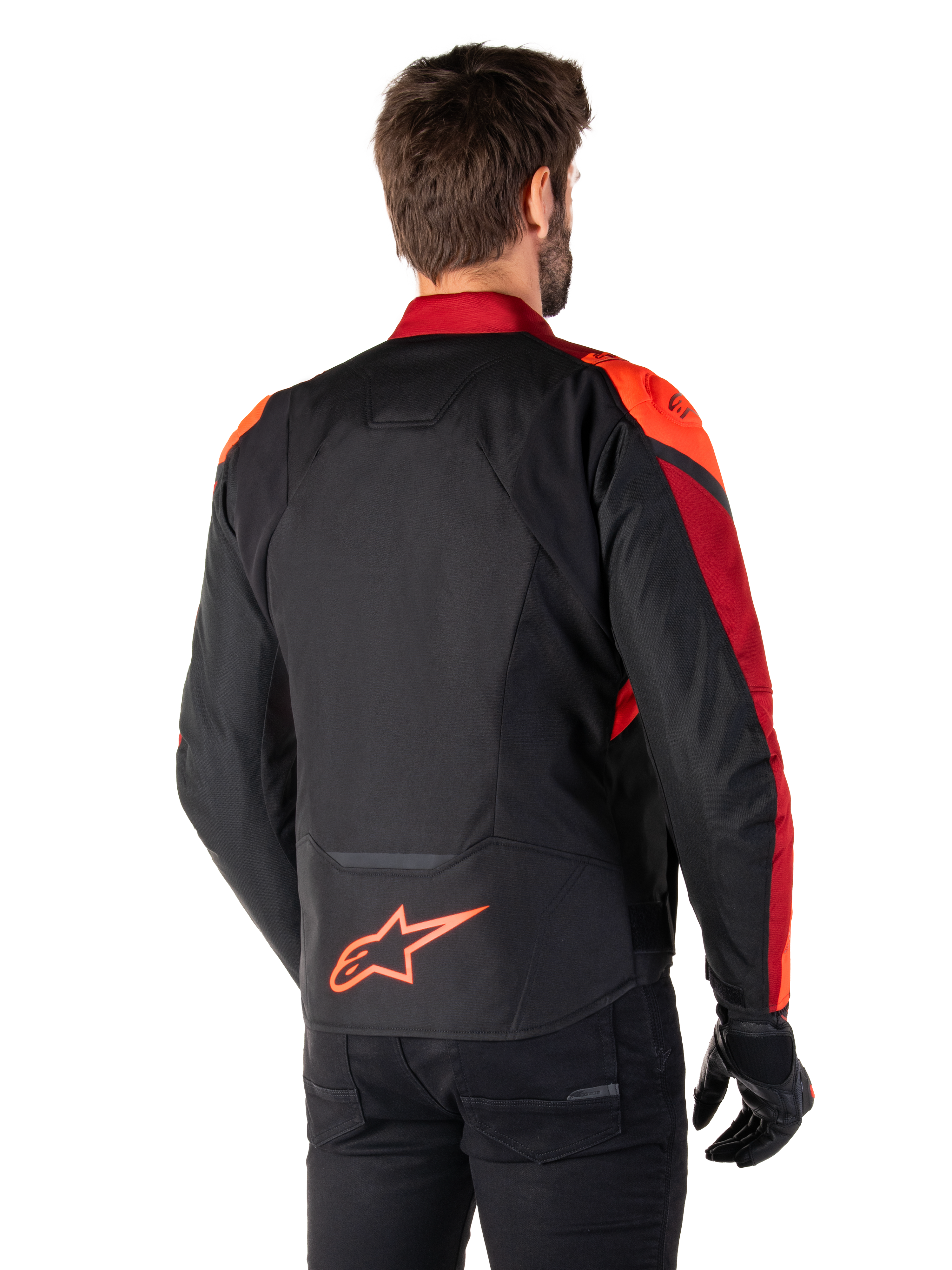 Vista trasera de un hombre con la chaqueta Alpinestars T-Jaws V4 Waterproof en Negro Rojo Fluo Rojo Oscuro, con detalles en rojo vibrante en hombros y mangas, logotipo de Alpinestars en rojo flúor en la parte inferior de la espalda, combinada con pantalones y guantes negros.