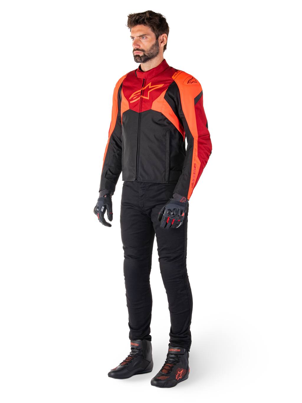 Piloto con la chaqueta Alpinestars T-Jaws V4 Waterproof en color negro, rojo flúor y rojo oscuro, combinada con pantalones de moto, guantes y botas de conducción negros, con un diseño textil de ajuste deportivo, logotipo llamativo en el pecho y una silueta agresiva.