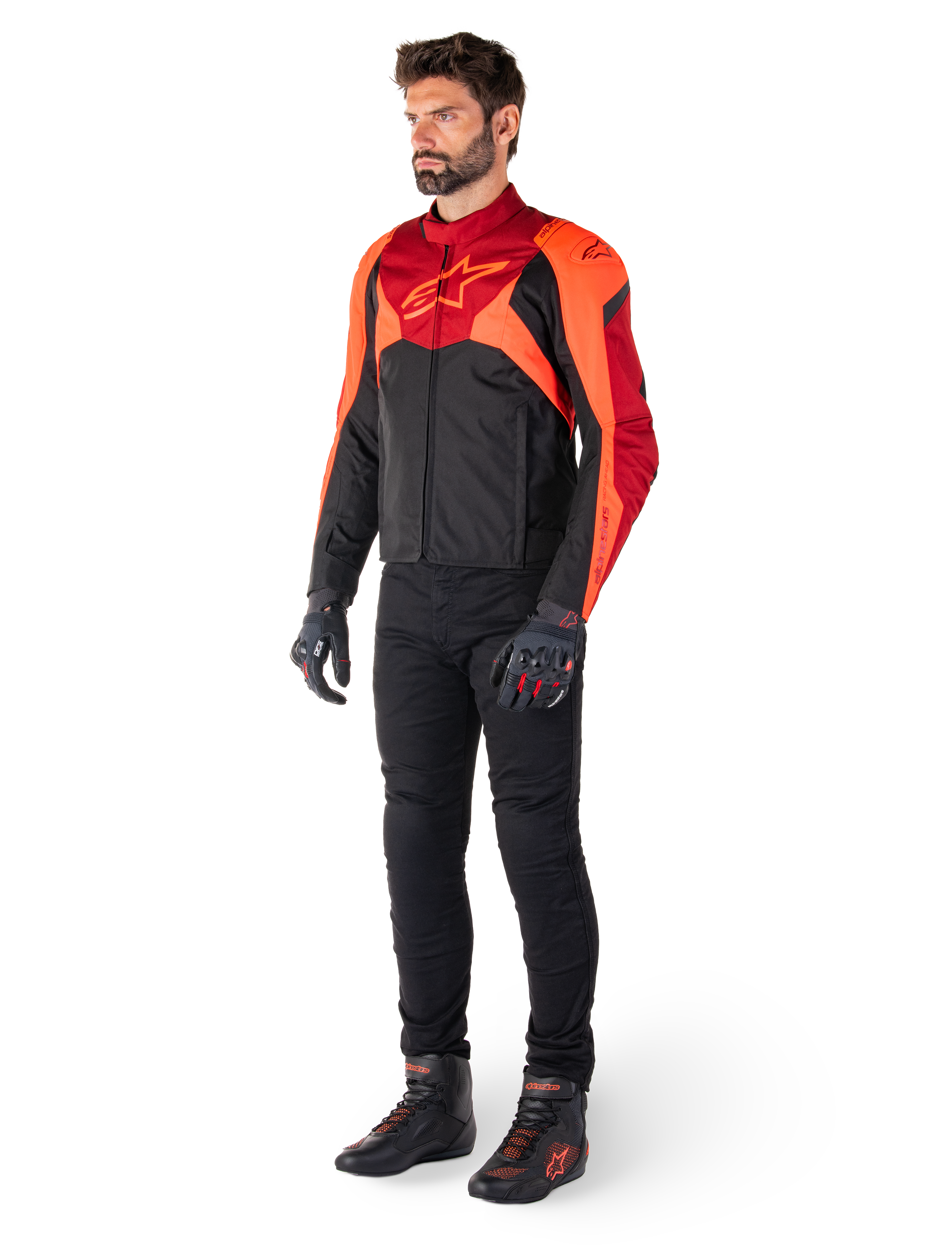 Piloto con la chaqueta Alpinestars T-Jaws V4 Waterproof en color negro, rojo flúor y rojo oscuro, combinada con pantalones de moto, guantes y botas de conducción negros, con un diseño textil de ajuste deportivo, logotipo llamativo en el pecho y una silueta agresiva.