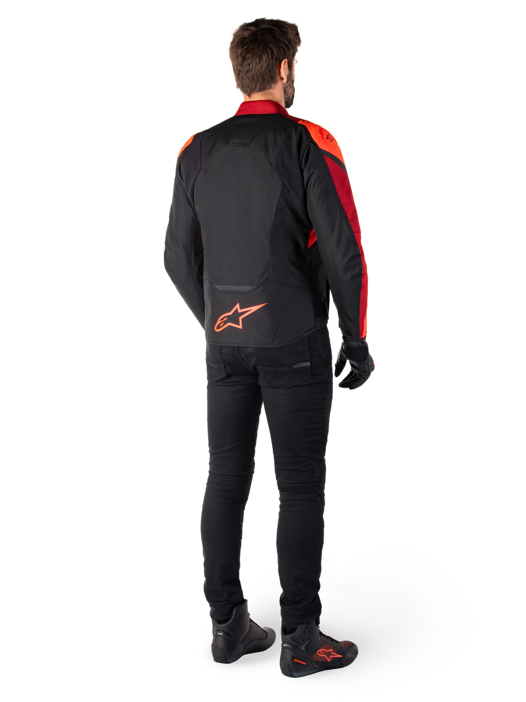 Chaqueta Alpinestars T-Jaws V4 Waterproof en color negro, rojo flúor y rojo oscuro, vista trasera de un piloto con la chaqueta textil, pantalones negros y botas de moto, con un llamativo logotipo rojo de Alpinestars en la parte inferior de la espalda.