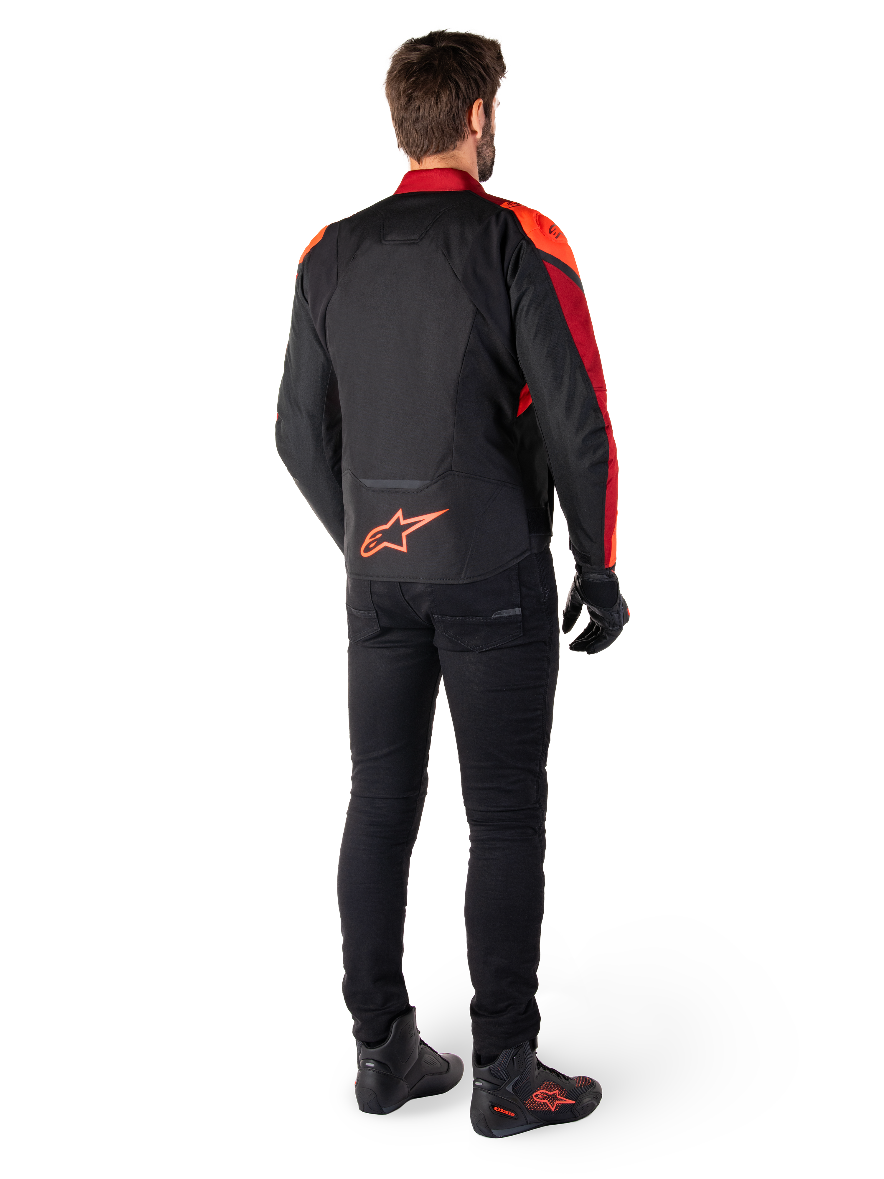 Chaqueta Alpinestars T-Jaws V4 Waterproof en color negro, rojo flúor y rojo oscuro, vista trasera de un piloto con la chaqueta textil, pantalones negros y botas de moto, con un llamativo logotipo rojo de Alpinestars en la parte inferior de la espalda.