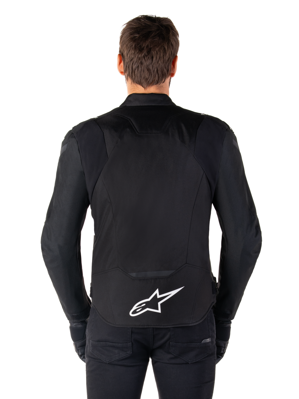 Chaqueta impermeable Alpinestars T-Jaws V4, chaqueta de moto negra vista desde atrás, con el logotipo blanco de Alpinestars en la parte inferior de la espalda, parte trasera extendida para mayor cobertura, usada por un modelo con pantalones y guantes negros, diseño textil deportivo y estilizado