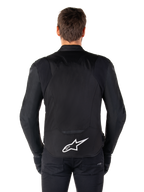 Chaqueta impermeable Alpinestars T-Jaws V4, chaqueta de moto negra vista desde atrás, con el logotipo blanco de Alpinestars en la parte inferior de la espalda, parte trasera extendida para mayor cobertura, usada por un modelo con pantalones y guantes negros, diseño textil deportivo y estilizado
