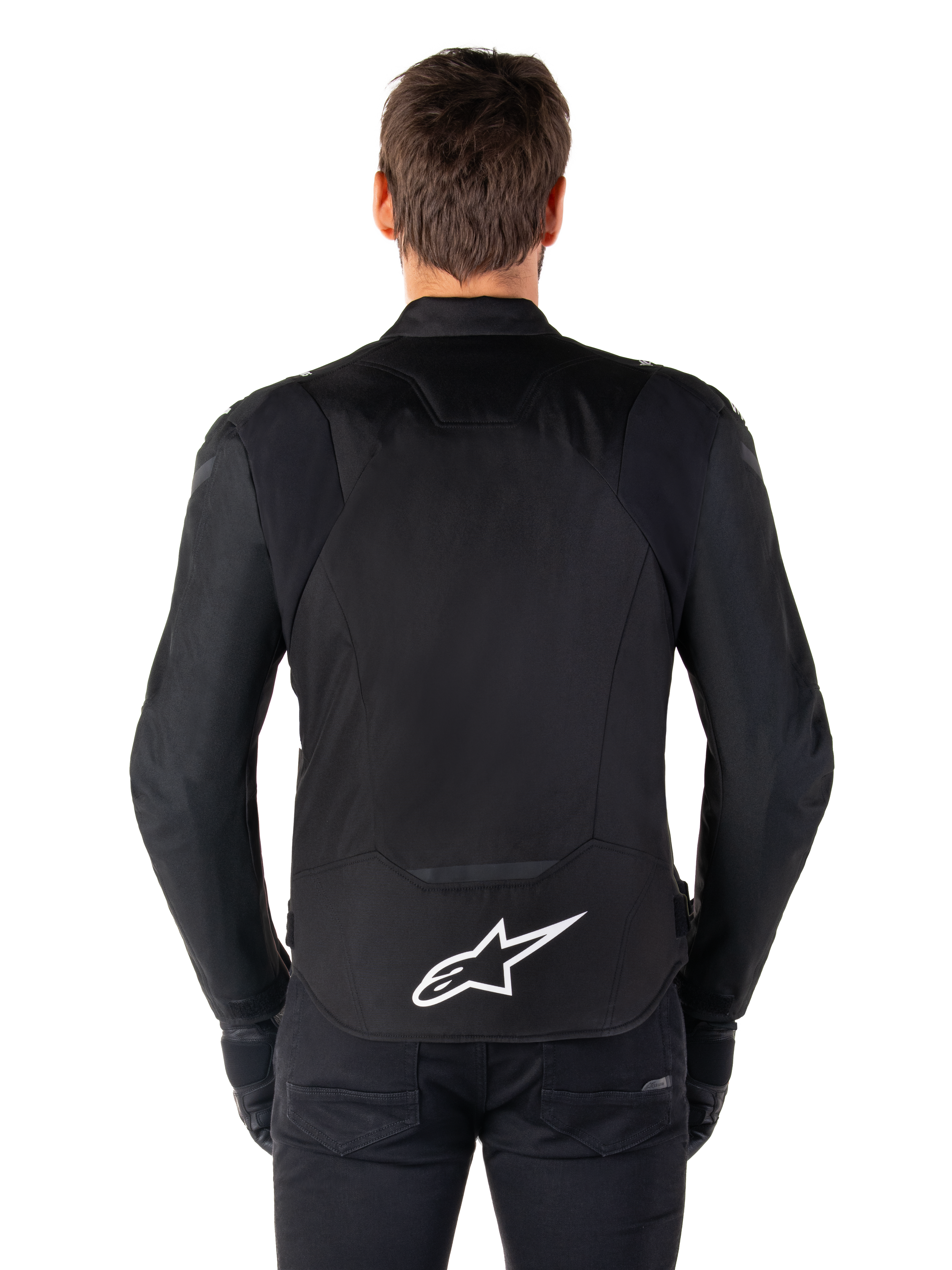 Chaqueta impermeable Alpinestars T-Jaws V4, chaqueta de moto negra vista desde atrás, con el logotipo blanco de Alpinestars en la parte inferior de la espalda, parte trasera extendida para mayor cobertura, usada por un modelo con pantalones y guantes negros, diseño textil deportivo y estilizado