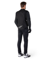 Chaqueta impermeable Alpinestars T-Jaws V4, negra y blanca, vista trasera que muestra el logotipo de la estrella blanca en la parte inferior de la espalda, usada por un modelo con pantalones de conducción negros, guantes negros y botas de moto blancas y negras.