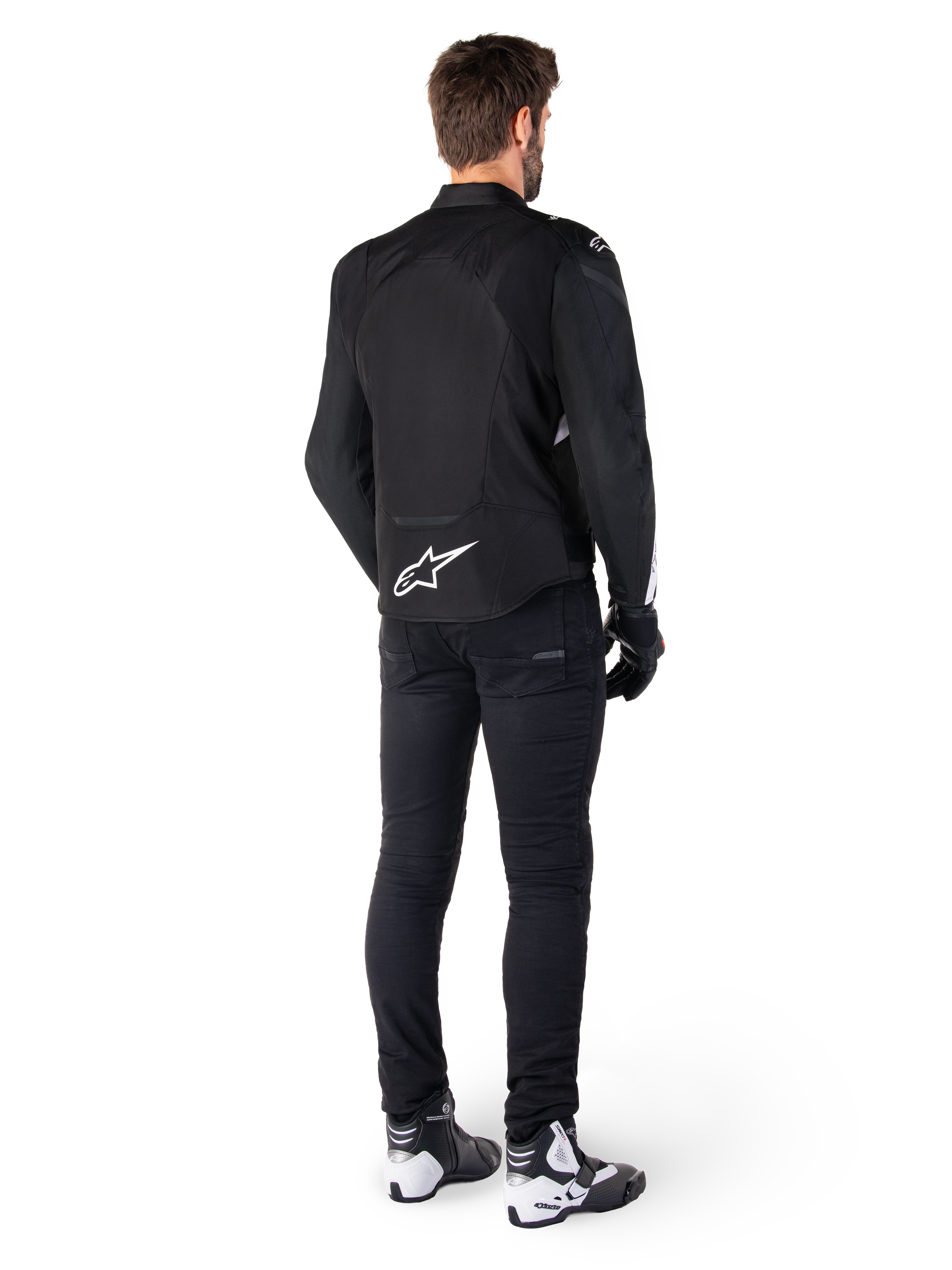 Chaqueta impermeable Alpinestars T-Jaws V4, negra y blanca, vista trasera que muestra el logotipo de la estrella blanca en la parte inferior de la espalda, usada por un modelo con pantalones de conducción negros, guantes negros y botas de moto blancas y negras.
