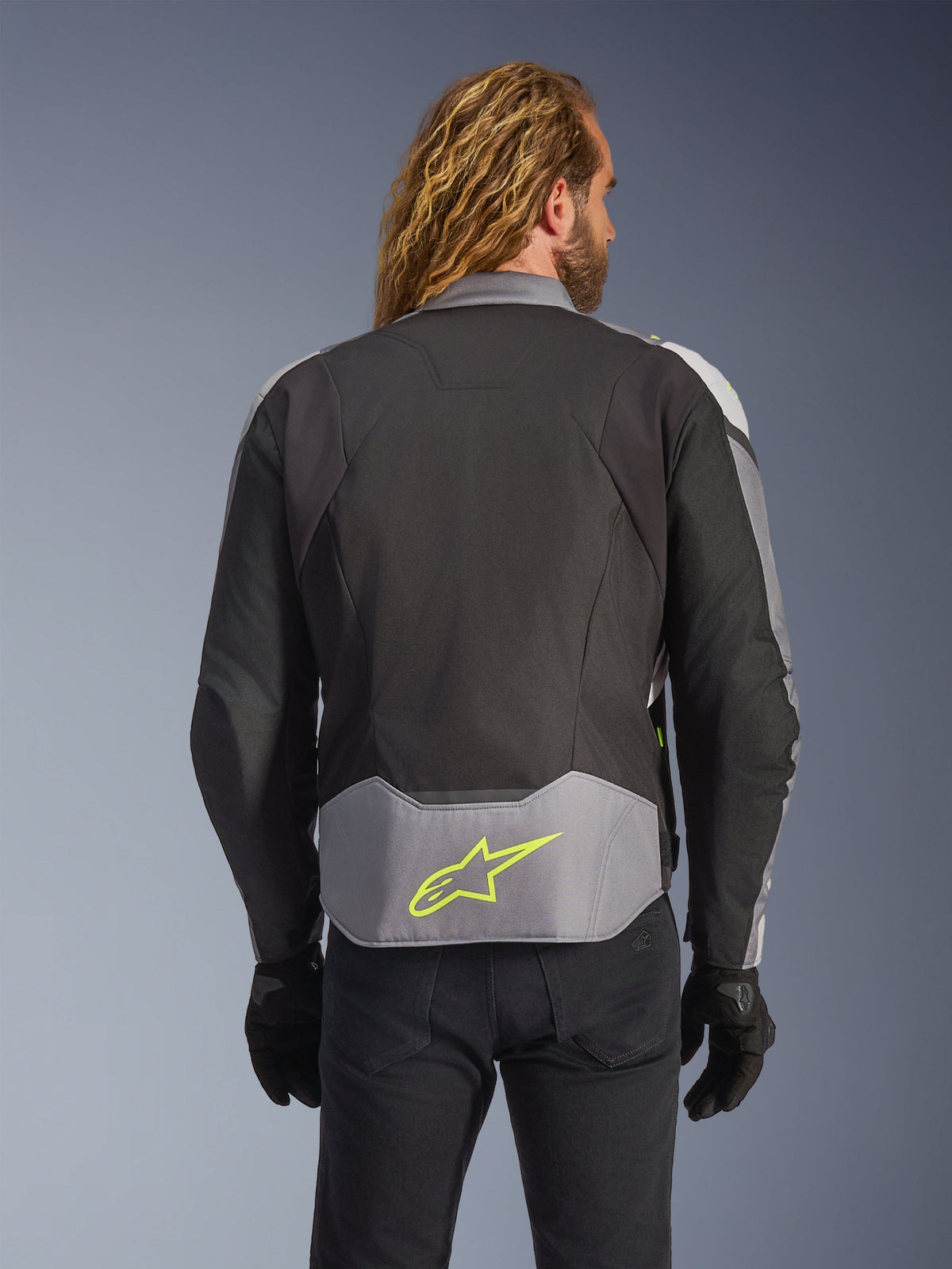 Chaqueta impermeable Alpinestars T-Jaws V4, color negro, gris hielo y gris oscuro, vista trasera en modelo, con un panel inferior en la espalda de color gris claro con un logotipo de estrella en amarillo neón prominente, construcción textil de múltiples paneles y silueta deportiva, combinada con guantes negros y pantalones oscuros.