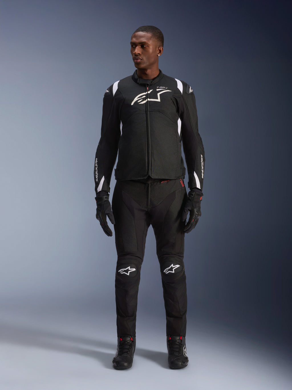 Chaqueta impermeable Alpinestars T-SP 1 V2, negra y blanca, con un logotipo blanco destacado en el pecho y detalles en los hombros, usada con pantalones de moto y guantes negros a juego para un look completo de conducción en carretera
