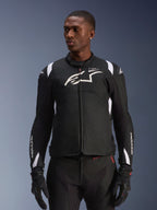 Chaqueta impermeable Alpinestars T-SP 1 V2, chaqueta textil deportiva para moto, en negro y blanco con sutiles detalles en rojo, usada por un modelo masculino con guantes y pantalones a juego, con el logotipo de Alpinestars en blanco en el pecho, marca T-SP-1 y silueta protectora para conducción en carretera
