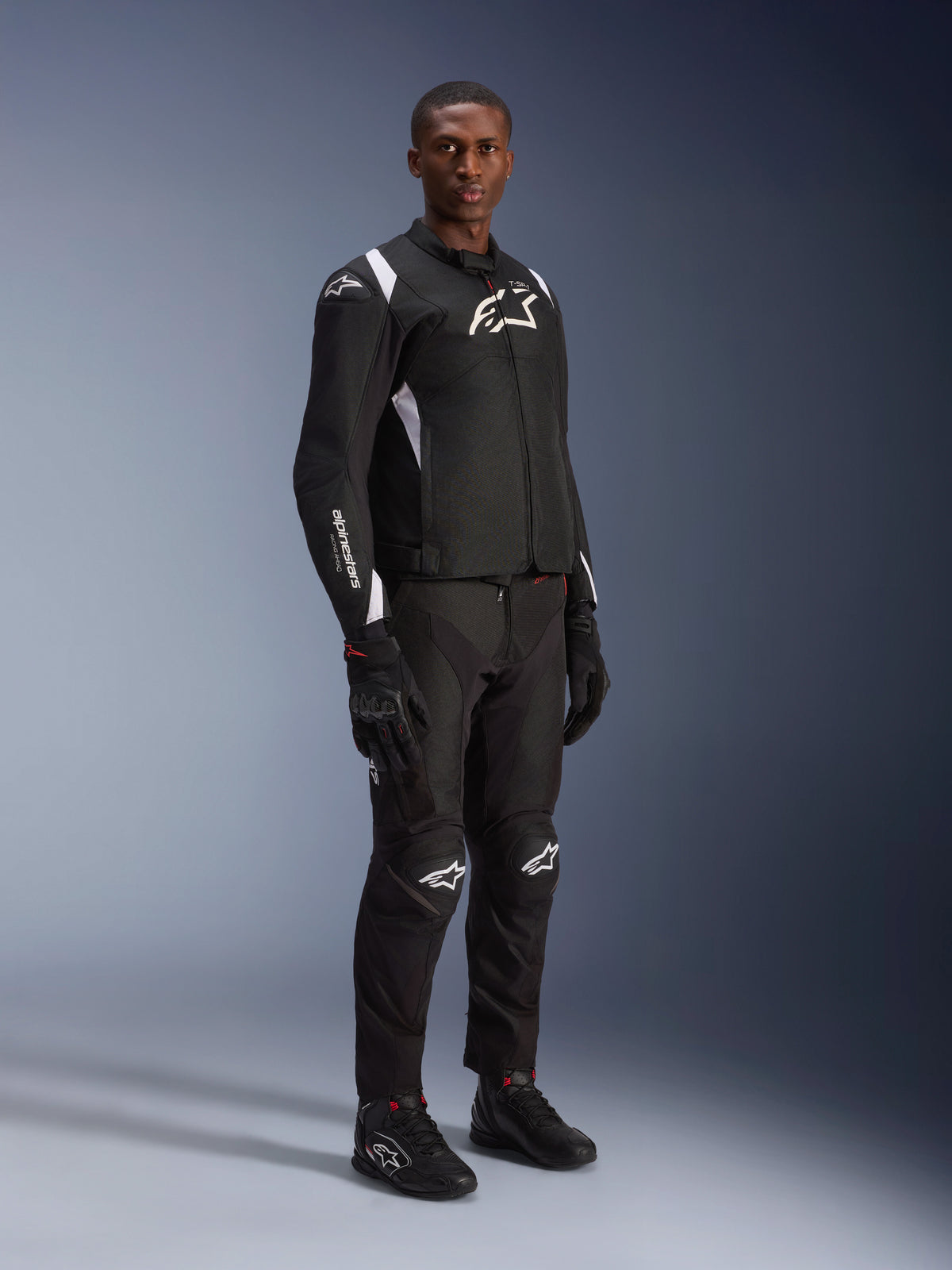Modelo masculino con la chaqueta impermeable Alpinestars T-SP 1 V2 en negro y blanco, combinada con pantalones de moto, guantes y botas negros a juego. Foto de estudio de cuerpo completo de una chaqueta textil deportiva con logotipos blancos y silueta entallada en 3D.