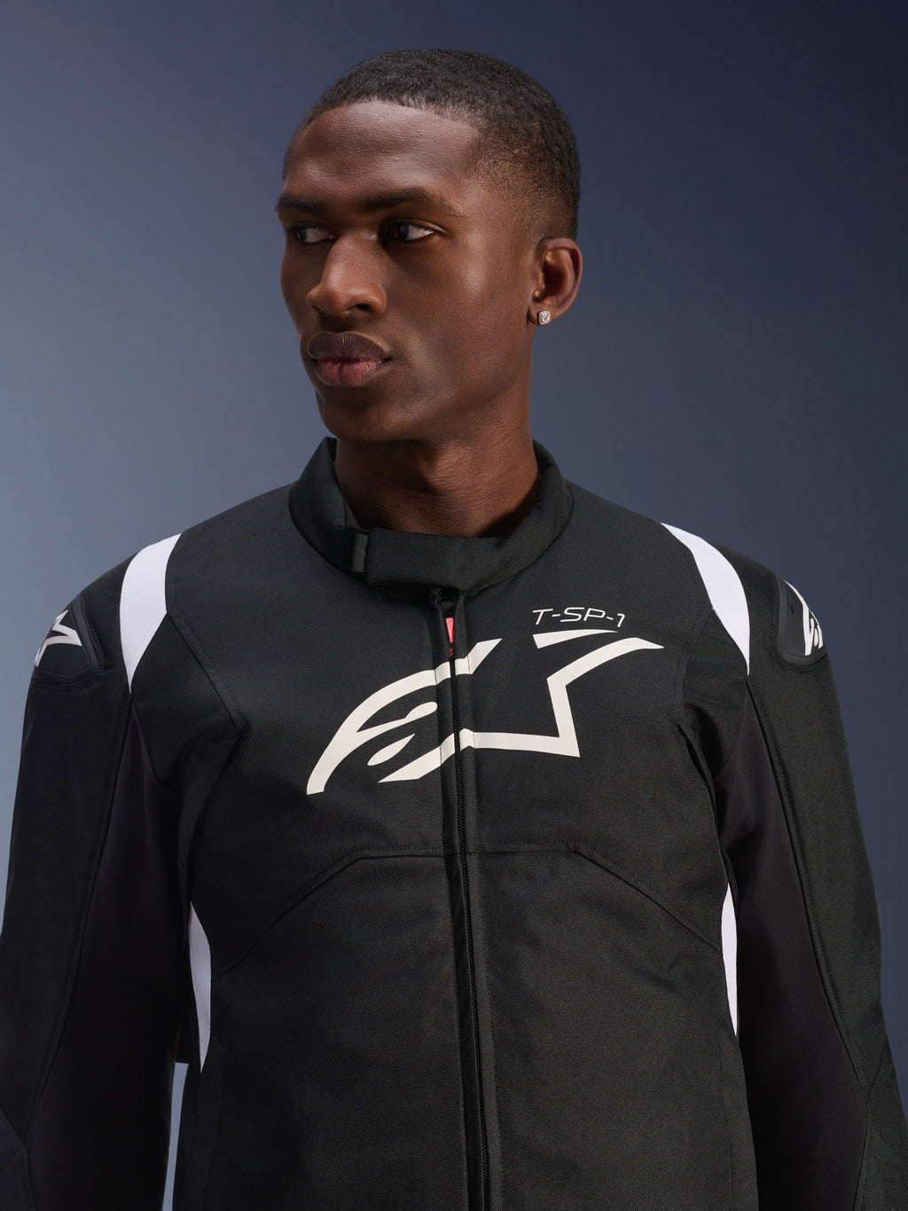 Chaqueta impermeable Alpinestars T-SP 1 V2 usada por un modelo, chaqueta textil de motociclismo en blanco y negro, logotipo grande de Alpinestars y marca T-SP-1 en el pecho, diseño deportivo con deslizadores protectores en los hombros