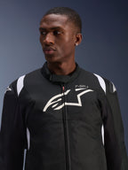 Chaqueta impermeable Alpinestars T-SP 1 V2 usada por un modelo, chaqueta textil de motociclismo en blanco y negro, logotipo grande de Alpinestars y marca T-SP-1 en el pecho, diseño deportivo con deslizadores protectores en los hombros