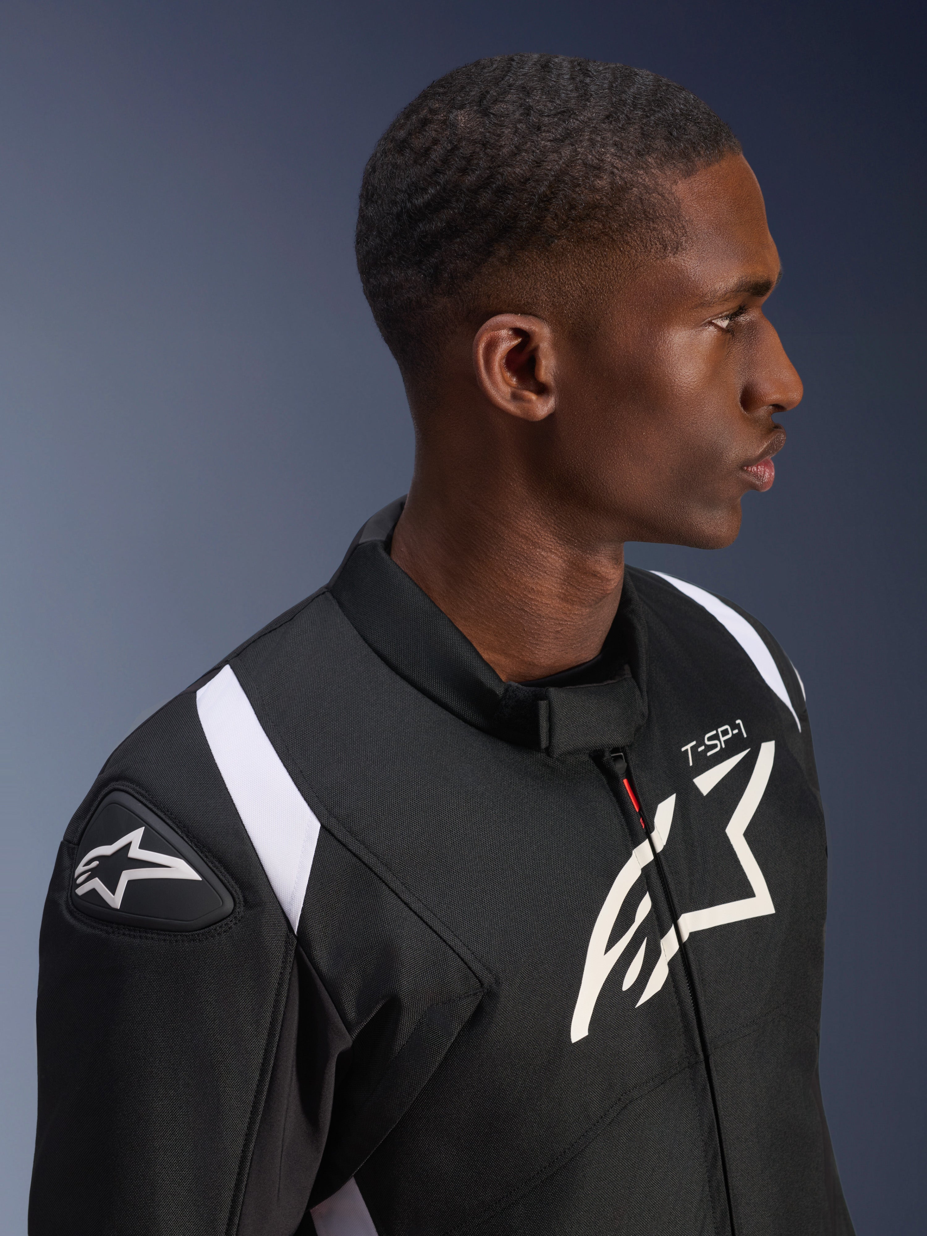 Chaqueta impermeable Alpinestars T-SP 1 V2 en negro y blanco, foto de perfil de un modelo masculino con la chaqueta puesta, destacando el logo de Alpinestars en el hombro y el pecho, la marca 'T-SP-1' y detalles blancos en los hombros.