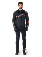 Modelo masculino con la chaqueta impermeable Alpinestars T-SPS V2 en color negro, blanco y rojo flúor, combinada con pantalones y guantes de moto negros, con logotipo de Alpinestars blanco en el pecho y detalles rojos en las mangas
