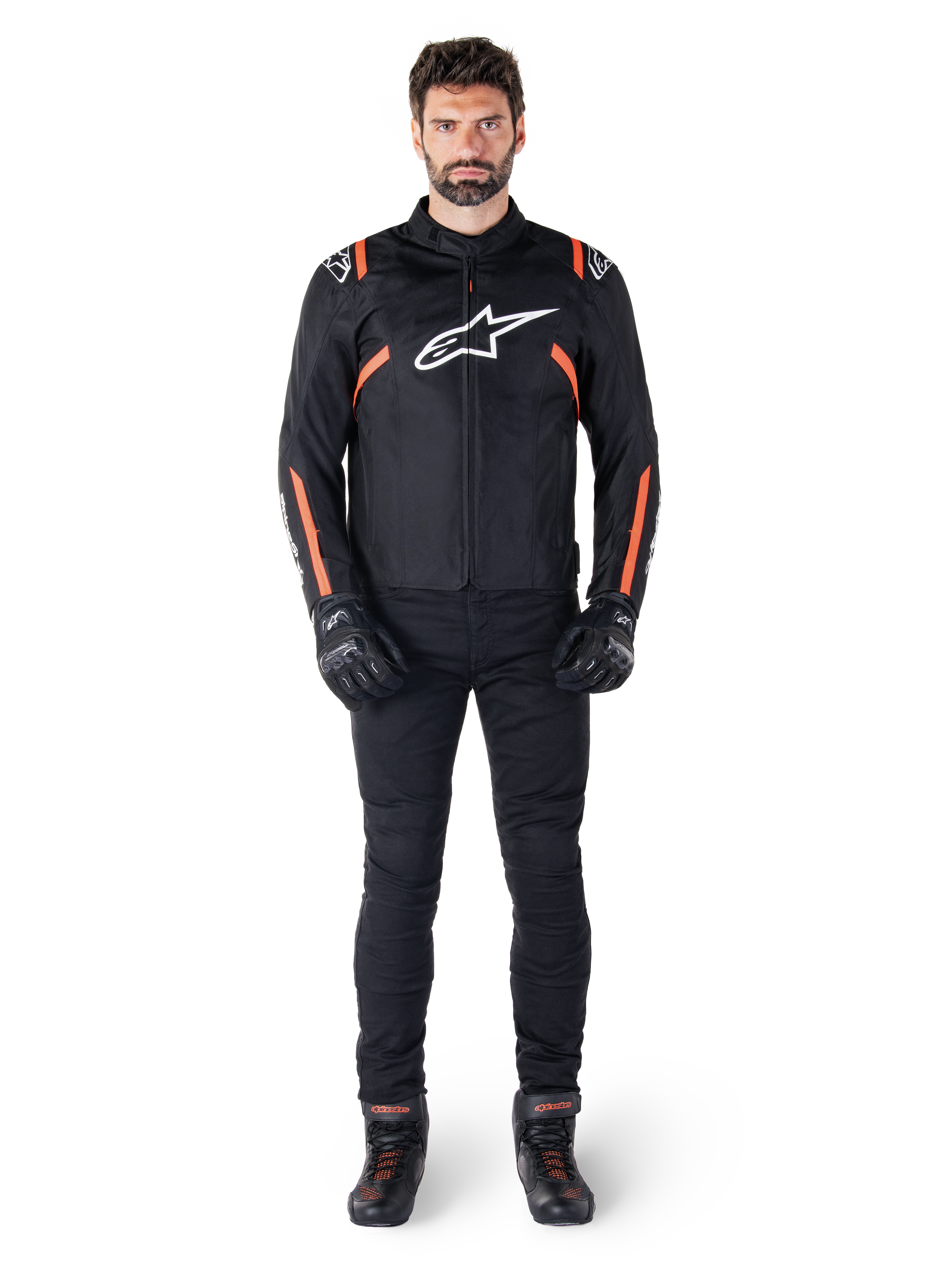 Modelo masculino con la chaqueta impermeable Alpinestars T-SPS V2 en color negro, blanco y rojo flúor, combinada con pantalones y guantes de moto negros, con logotipo de Alpinestars blanco en el pecho y detalles rojos en las mangas