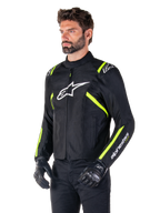 Modelo masculino con la chaqueta Alpinestars T-SPS V2 Waterproof, negra con detalles en blanco y amarillo flúor, con el logotipo Astars blanco en el pecho y franjas amarillo flúor en hombros y mangas, combinada con pantalones negros y guantes de conducción, chaqueta textil de moto de corte deportivo.