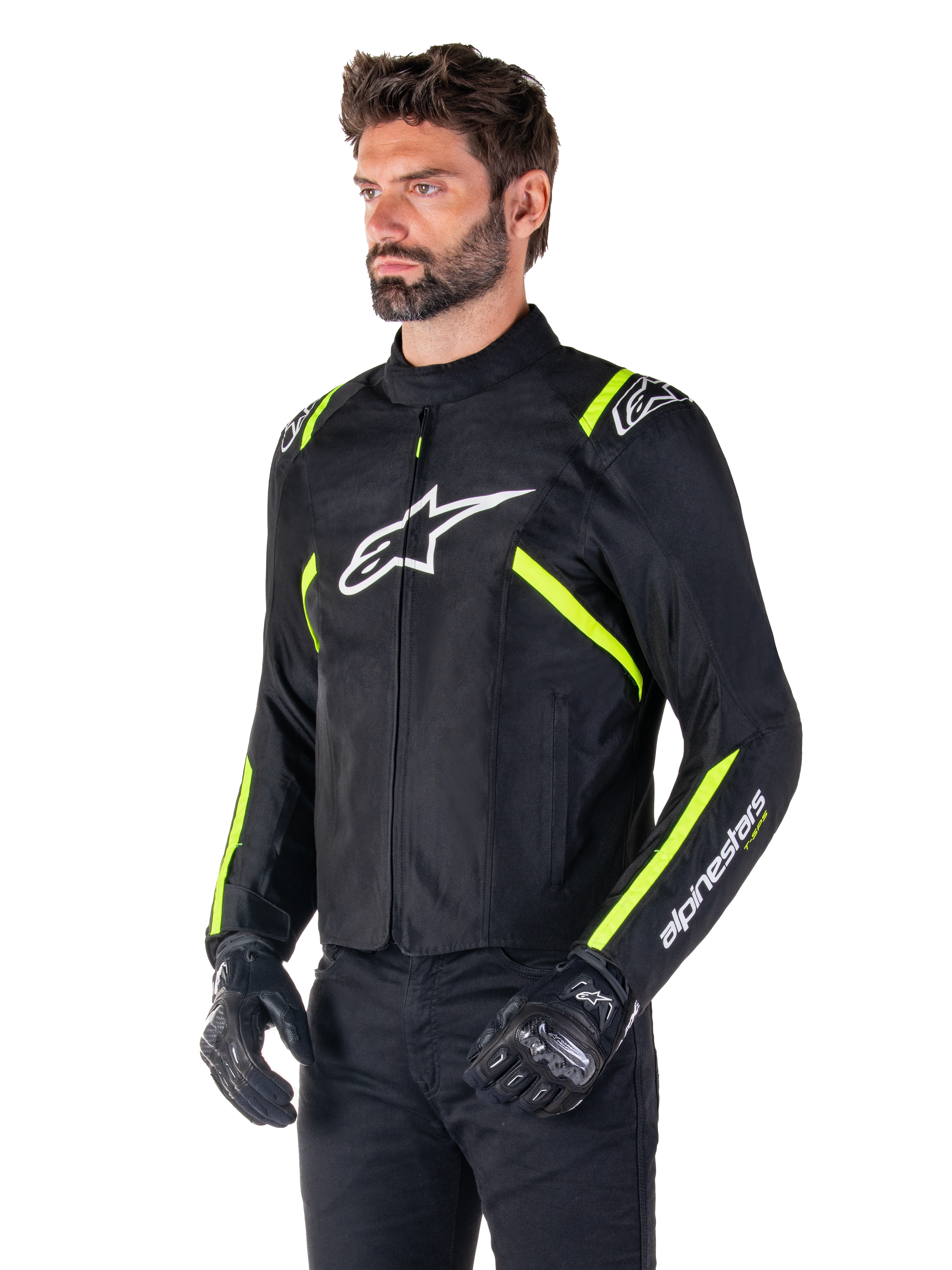 Modelo masculino con la chaqueta Alpinestars T-SPS V2 Waterproof, negra con detalles en blanco y amarillo flúor, con el logotipo Astars blanco en el pecho y franjas amarillo flúor en hombros y mangas, combinada con pantalones negros y guantes de conducción, chaqueta textil de moto de corte deportivo.