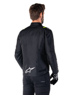 Chaqueta impermeable Alpinestars T-SPS V2, Negro Blanco Amarillo Flúor, vista trasera de modelo masculino vistiendo la chaqueta textil de moto, con un logotipo blanco en la parte inferior de la espalda y detalles en amarillo flúor en los hombros, combinada con pantalones y guantes negros.