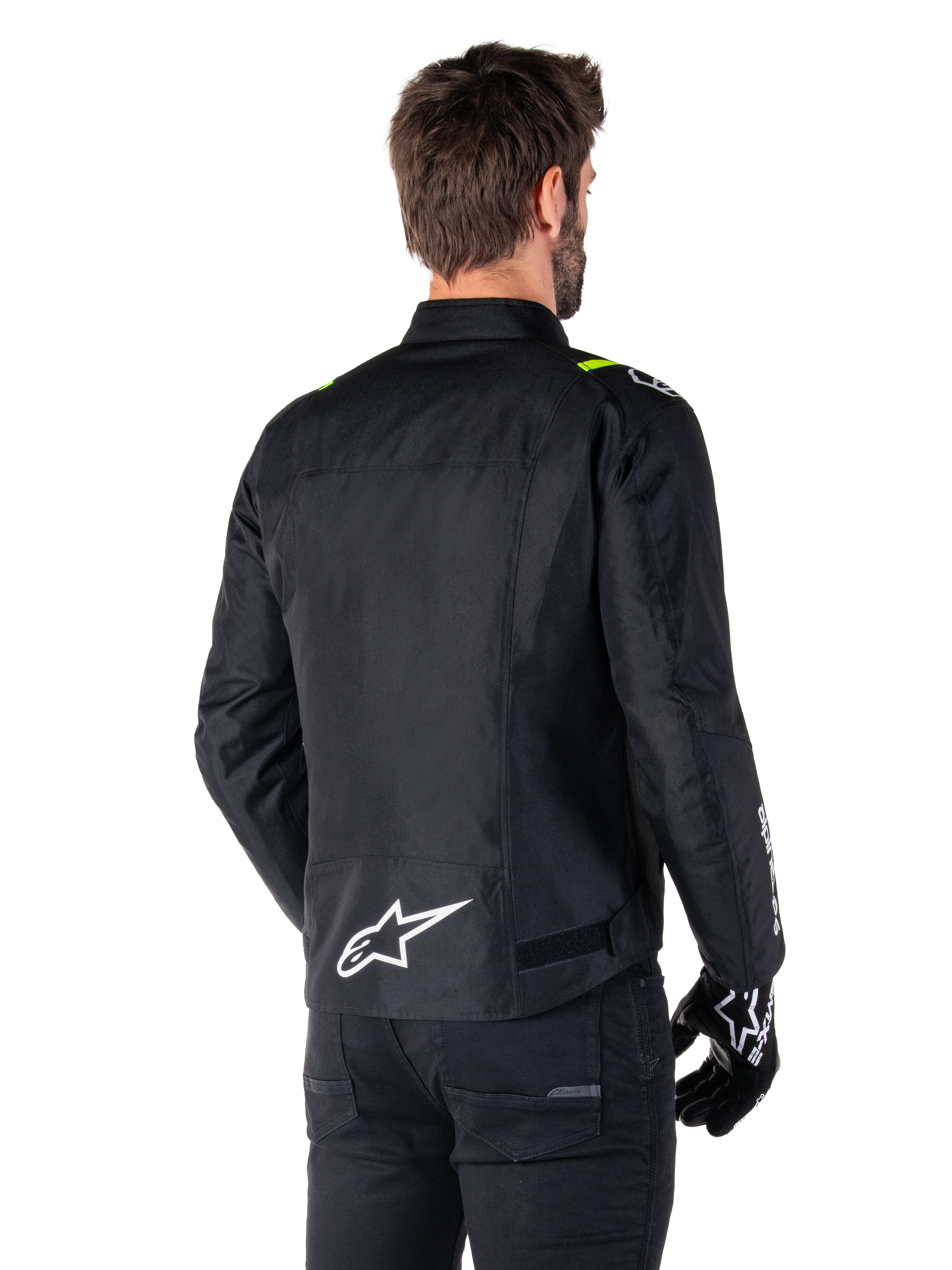 Chaqueta impermeable Alpinestars T-SPS V2, Negro Blanco Amarillo Flúor, vista trasera de modelo masculino vistiendo la chaqueta textil de moto, con un logotipo blanco en la parte inferior de la espalda y detalles en amarillo flúor en los hombros, combinada con pantalones y guantes negros.