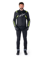 Chaqueta impermeable Alpinestars T-SPS V2, negro, blanco y amarillo flúor, lucida por un modelo con pantalones de moto y guantes negros, con logotipo de Alpinestars blanco en el pecho y detalles fluorescentes en hombros y mangas, equipamiento de motociclismo de corte deportivo
