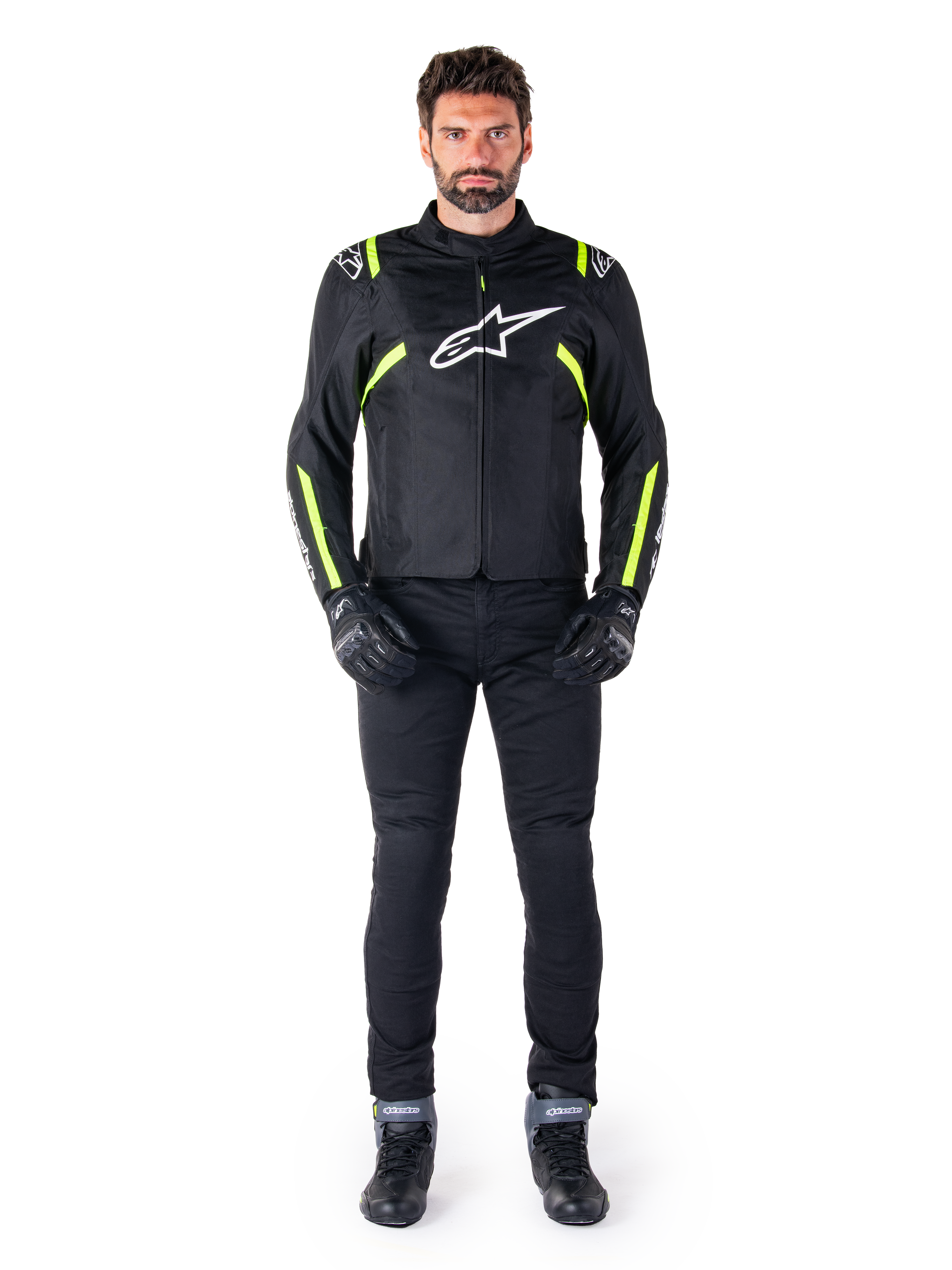 Chaqueta impermeable Alpinestars T-SPS V2, negro, blanco y amarillo flúor, lucida por un modelo con pantalones de moto y guantes negros, con logotipo de Alpinestars blanco en el pecho y detalles fluorescentes en hombros y mangas, equipamiento de motociclismo de corte deportivo