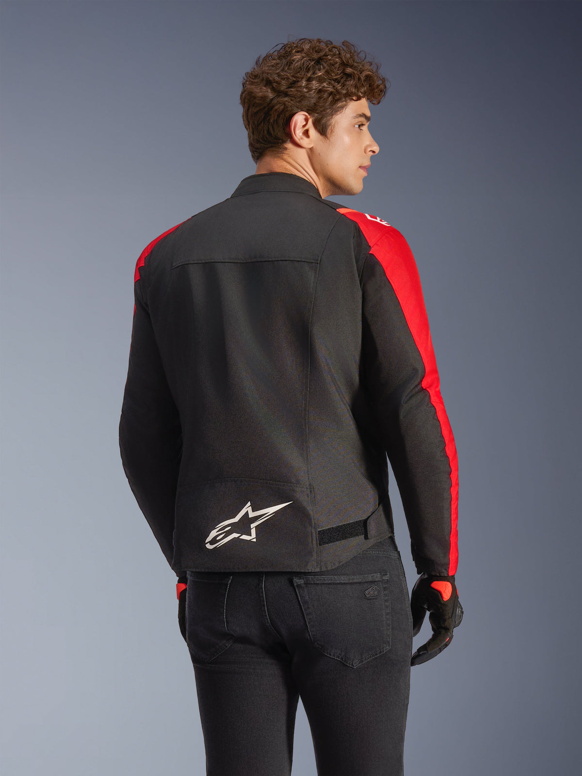 Chaqueta impermeable Alpinestars T-SPS V2 en negro y rojo, vista posterior en un modelo, con un logotipo blanco de Alpinestars en la parte inferior de la espalda, paneles vibrantes en rojo en las mangas y silueta de corte deportivo, usada con guantes negros y vaqueros.
