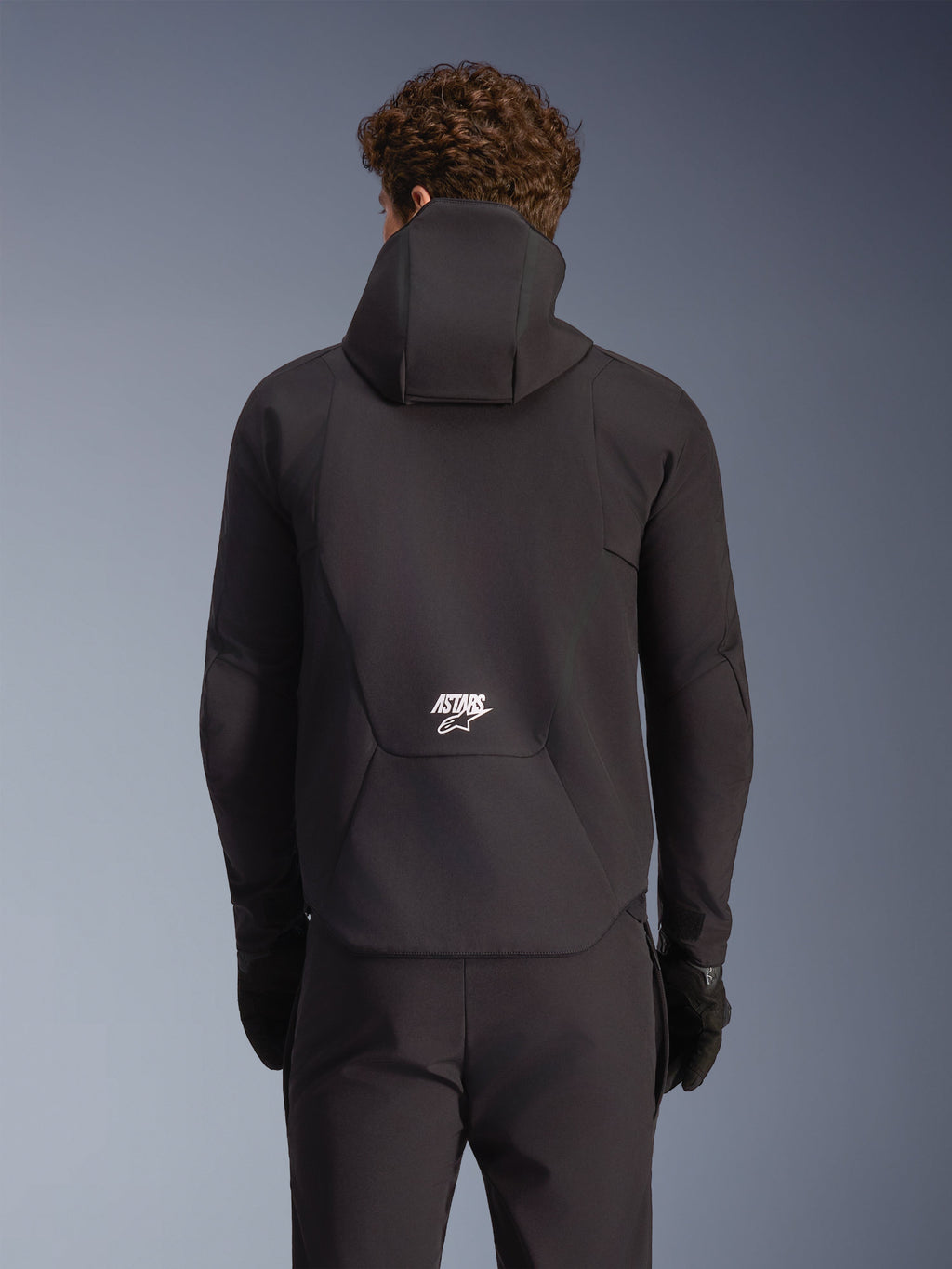 Alpinestars Aeroshell impermeable, chaqueta de moto softshell negra, vista trasera en modelo con capucha y logo blanco.