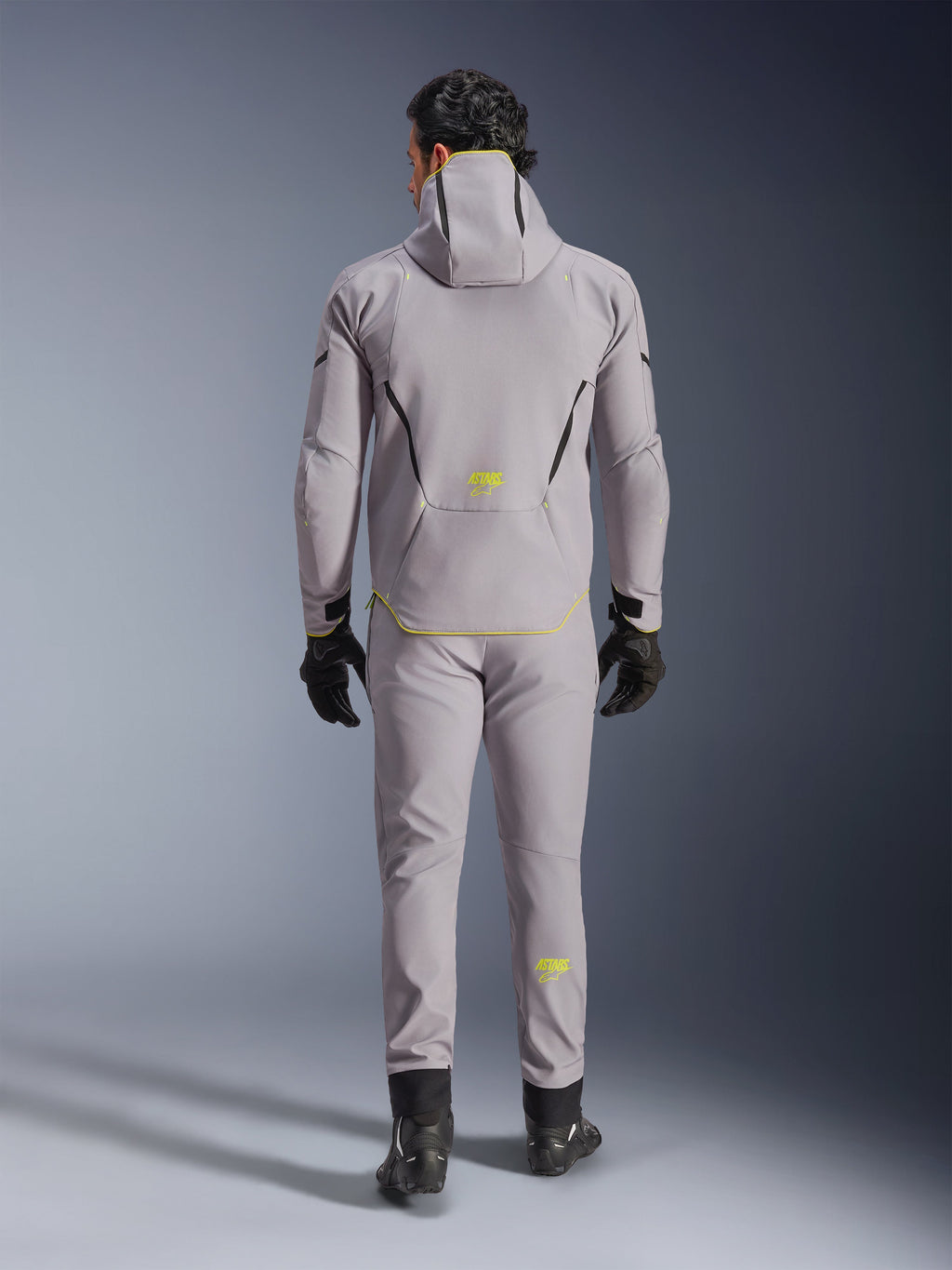 Alpinestars Aeroshell Waterproof Jacket in Mid Gray Yellow Fluo, full back view, worn with matching gray pants, black gloves, and black riding shoes. Features a hooded softshell design with yellow fluo 'ASTARS' branding and accents, engineered for modern motorcycle road performance.